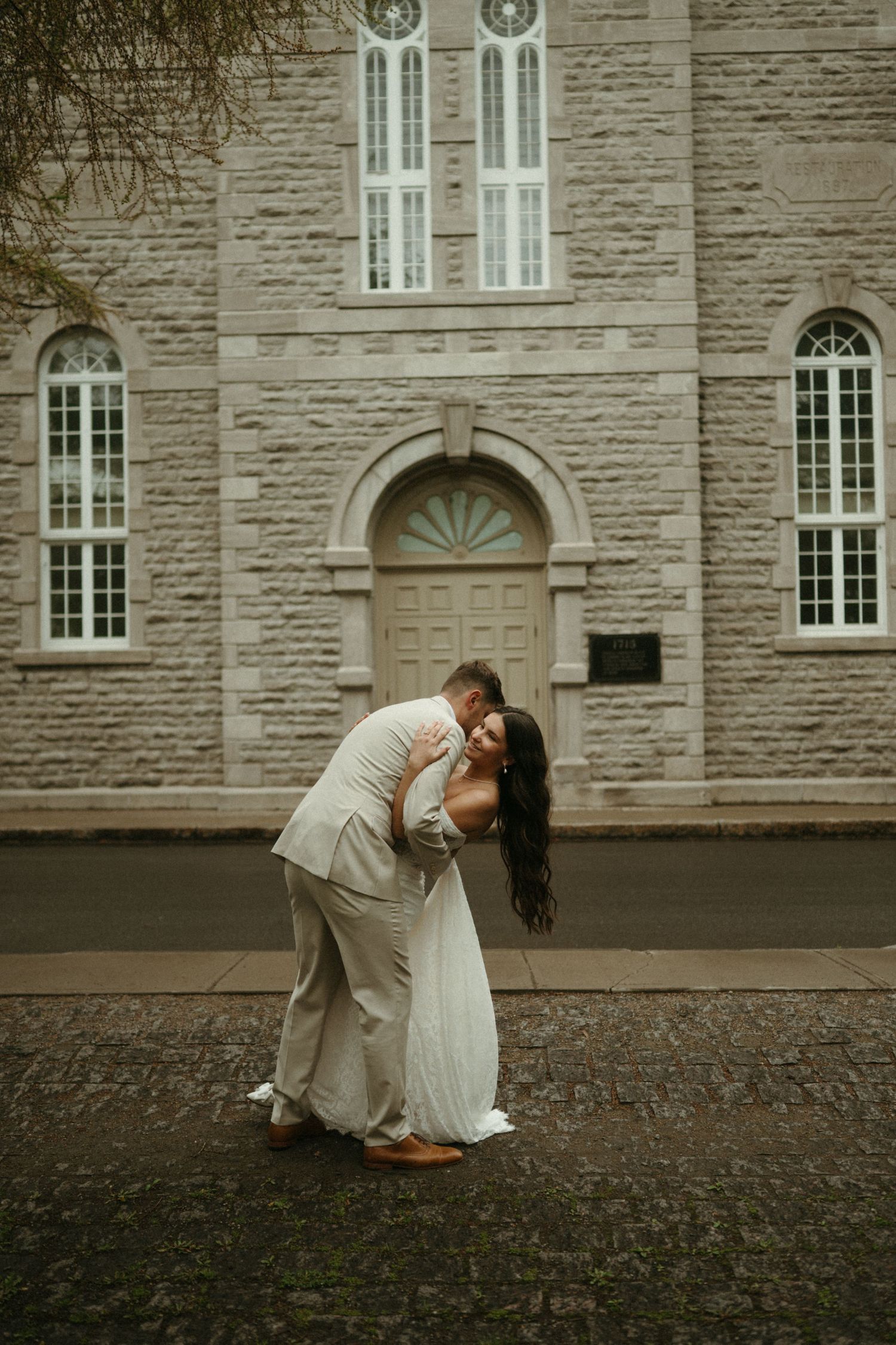 Wedding couple embracing in a dip pose before elegant stone church entrance on overcast day.