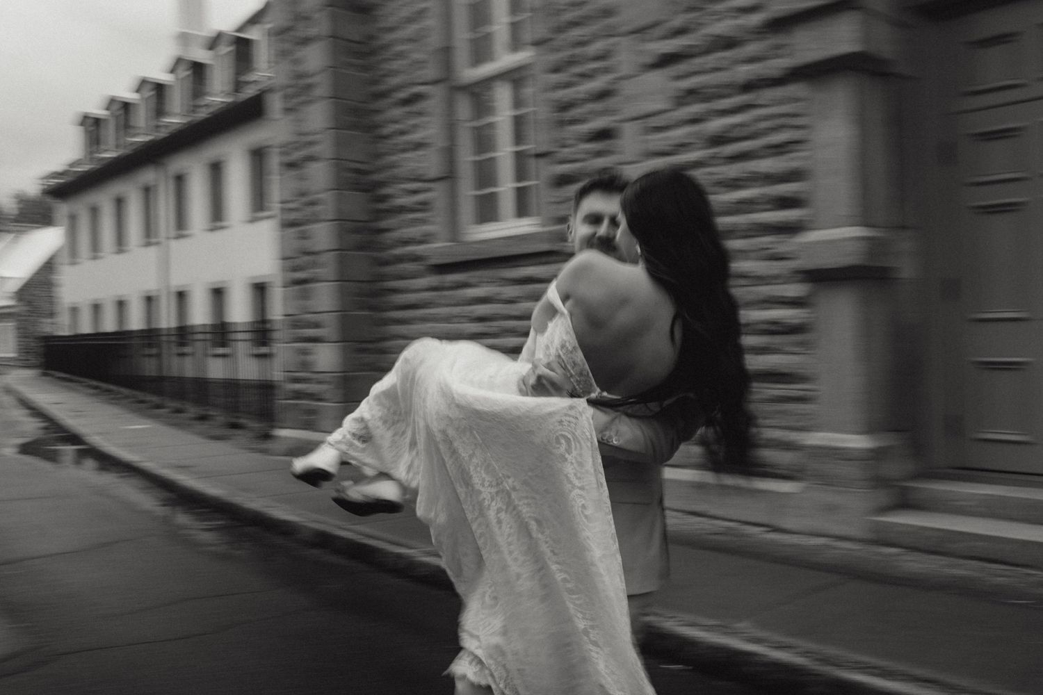 Groom carries bride in white wedding dress through cobblestone street in romantic black and white photo.