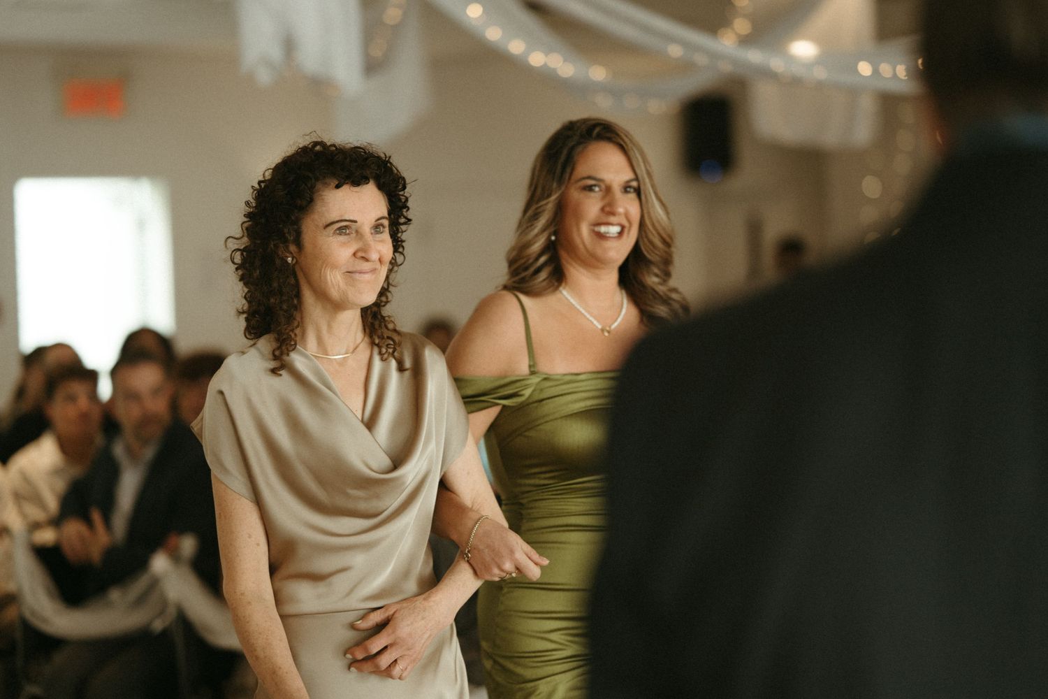 Two women smiling and walking arm-in-arm at a wedding reception in a decorated venue.