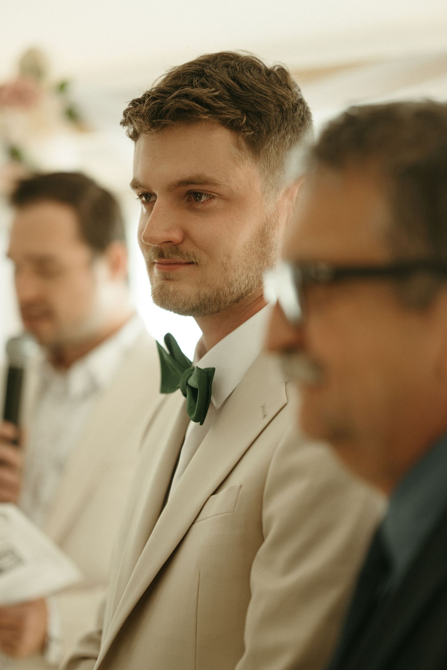 Groom in beige suit with green bow tie listening attentively during wedding ceremony.