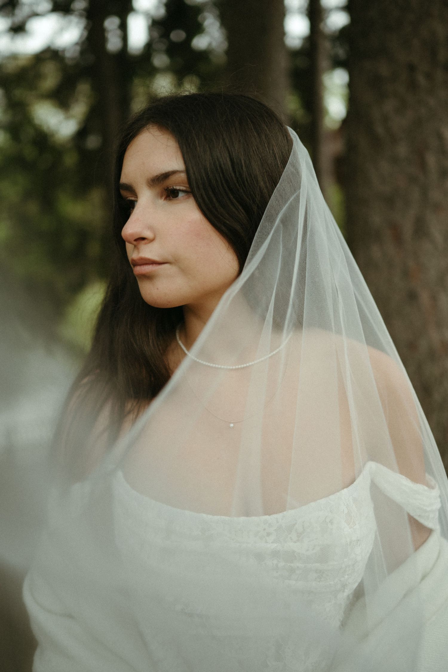 Bride in white off-shoulder gown and flowing veil gazes thoughtfully among trees in an ethereal outdoor portrait.