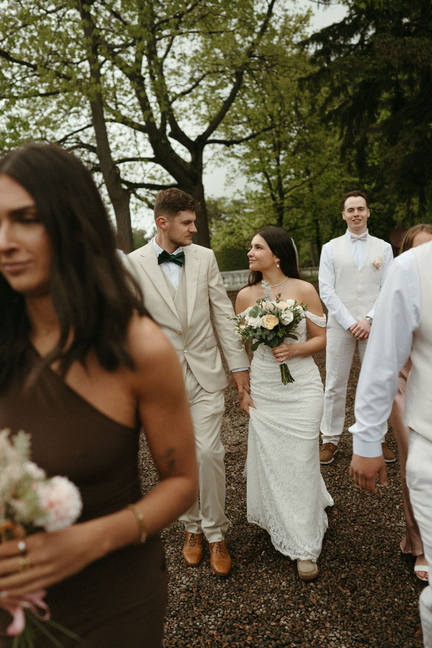 Bride and groom walk with wedding party on gravel path surrounded by lush green trees.