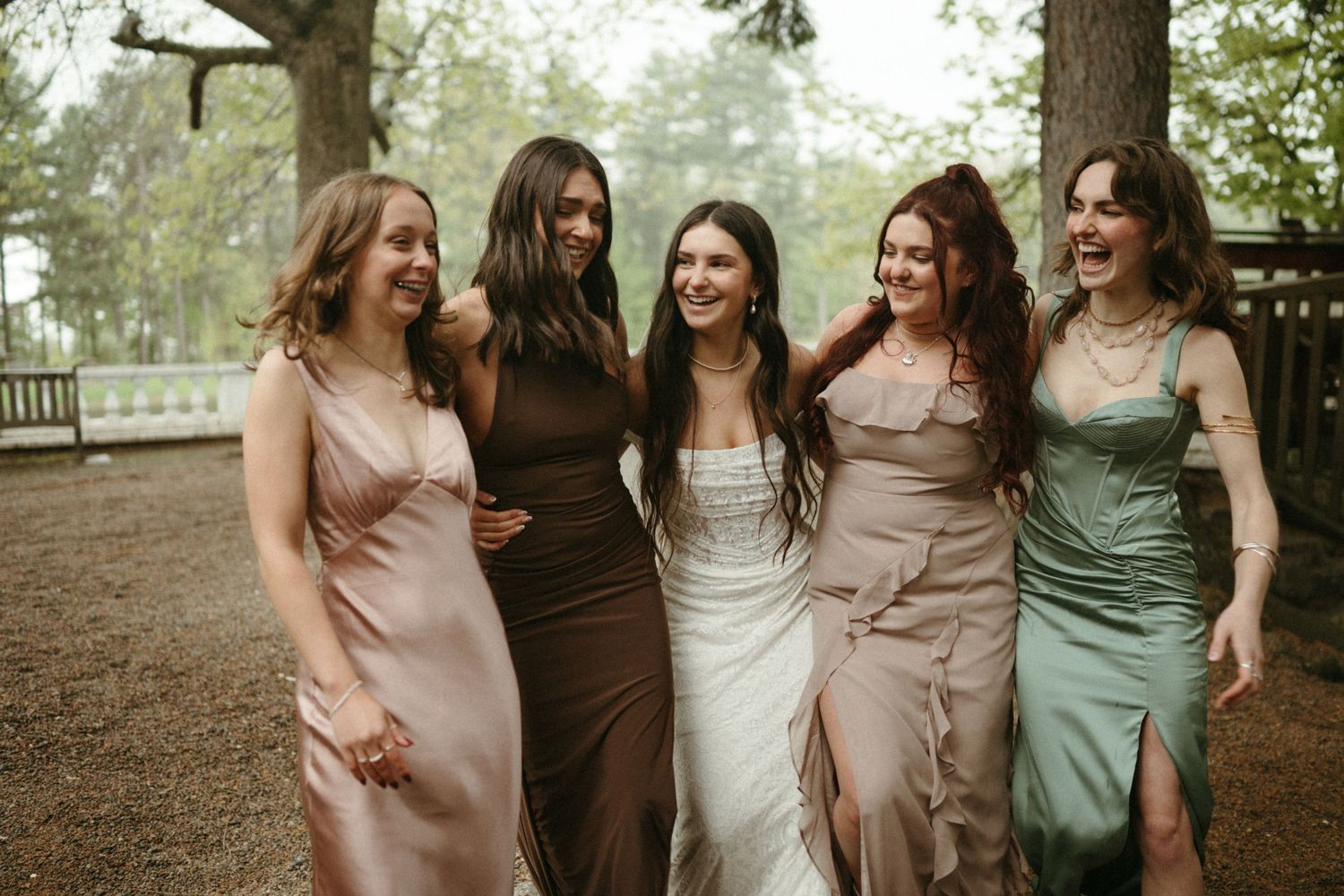 Bride in white gown laughing with four bridesmaids in mismatched satin dresses under trees outdoors.