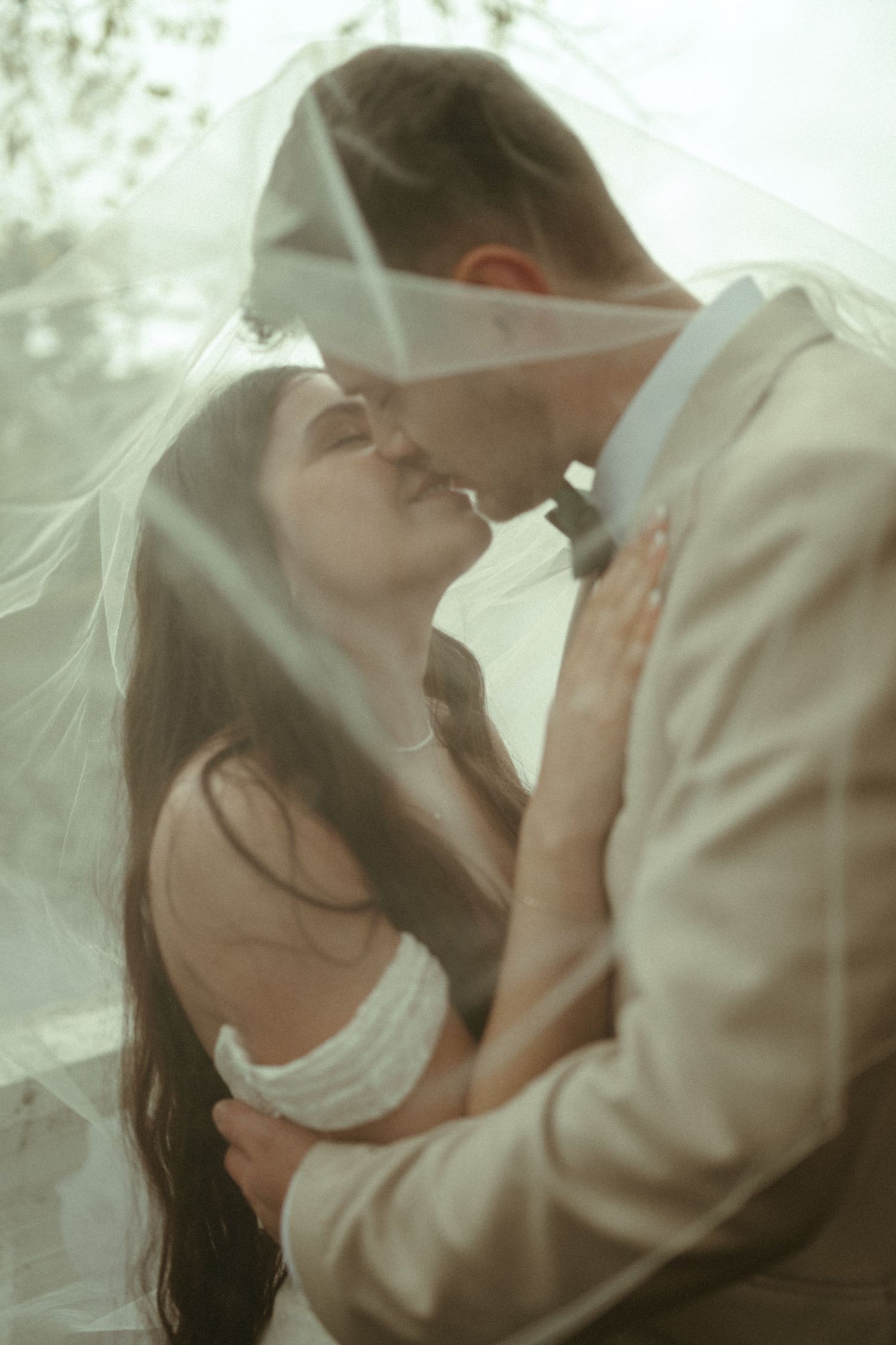 Bride and groom share a kiss under a flowing white veil in a soft, dreamy outdoor setting.