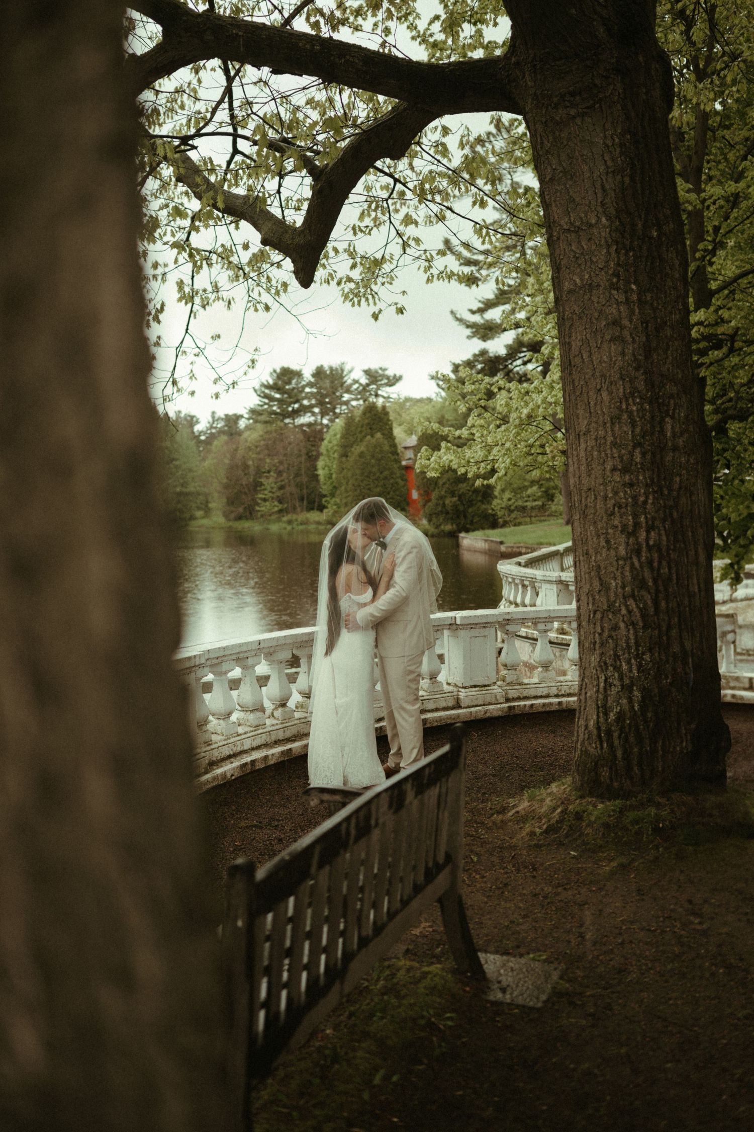 Bride and groom kissing on white balustrade bridge surrounded by lush trees beside serene lake.