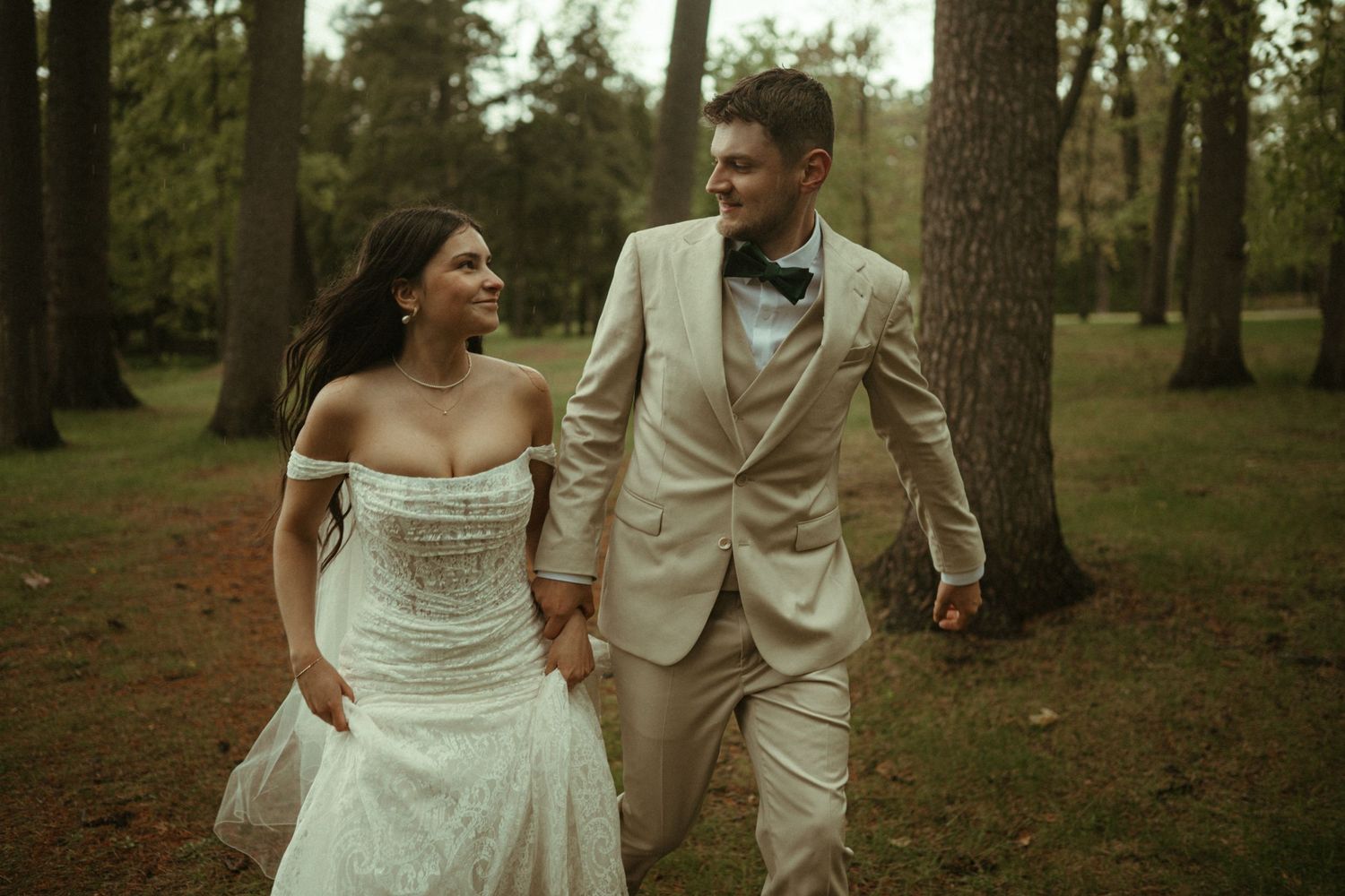 Smiling bride and groom holding hands walking through a forest in wedding attire.