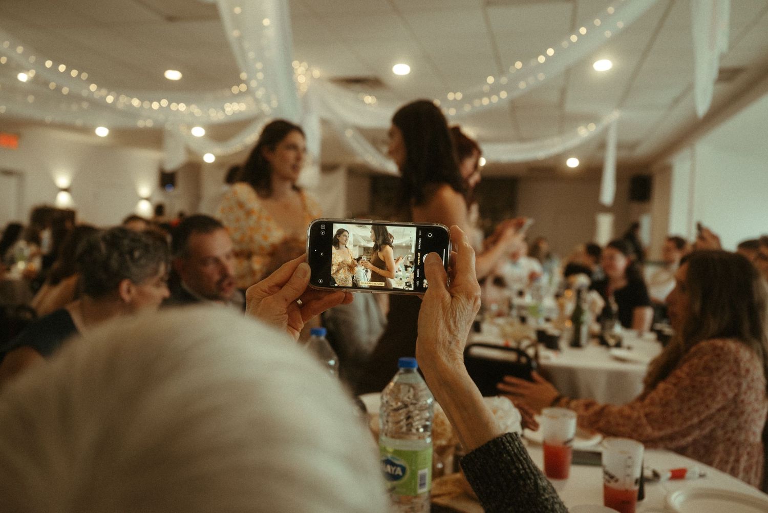 Wedding guest capturing bride on smartphone during lively indoor reception celebration.