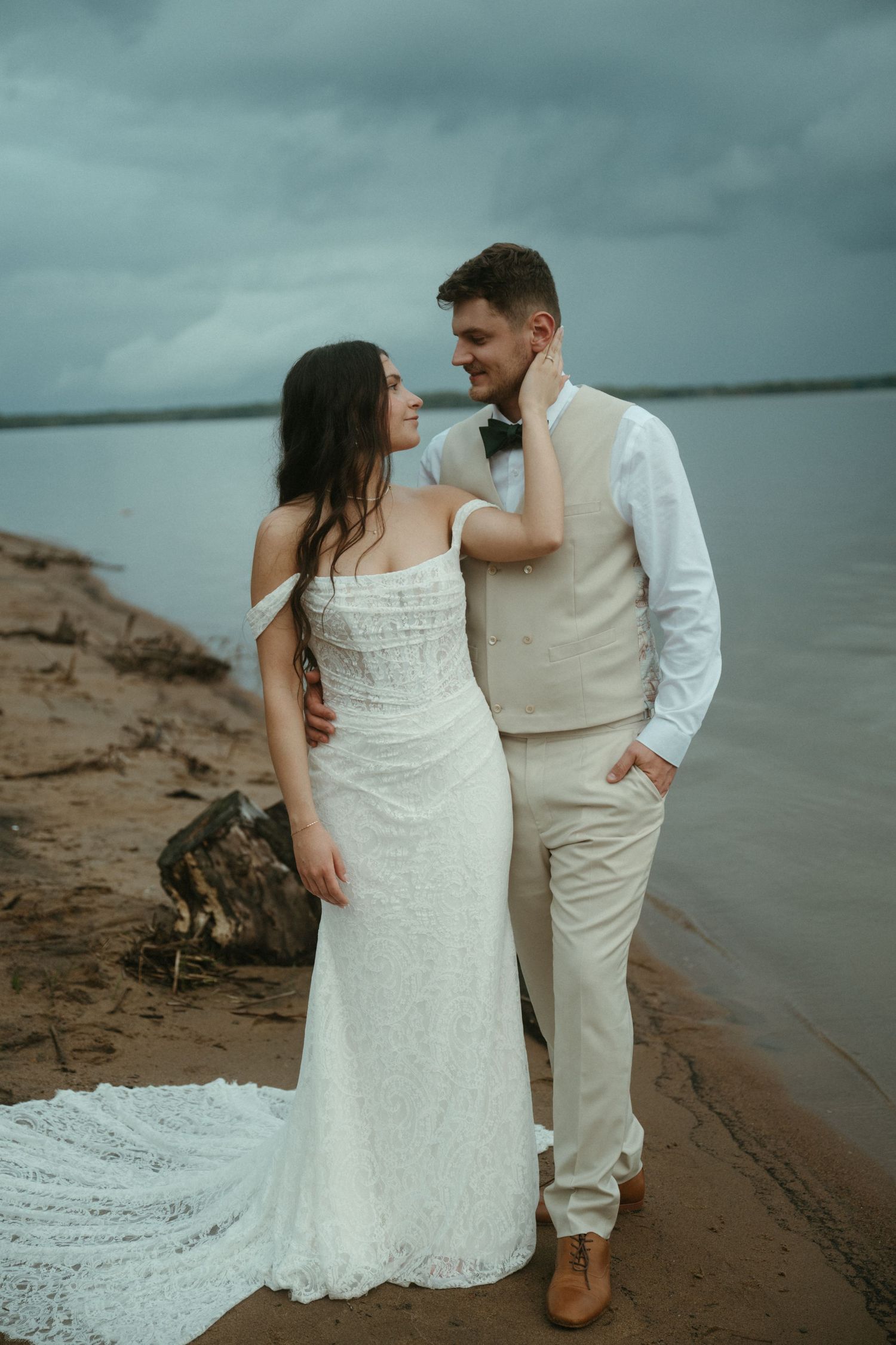 Bride and groom share a romantic moment on a sandy beach shoreline, with the bride in a white lace gown.