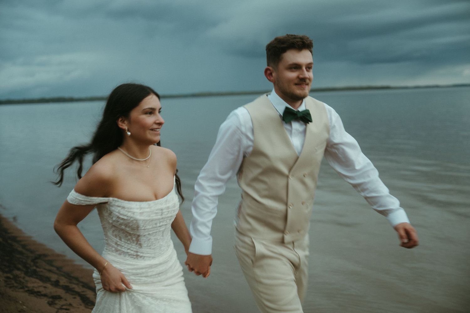 Bride and groom holding hands running along a lakeside shore under dramatic stormy skies.
