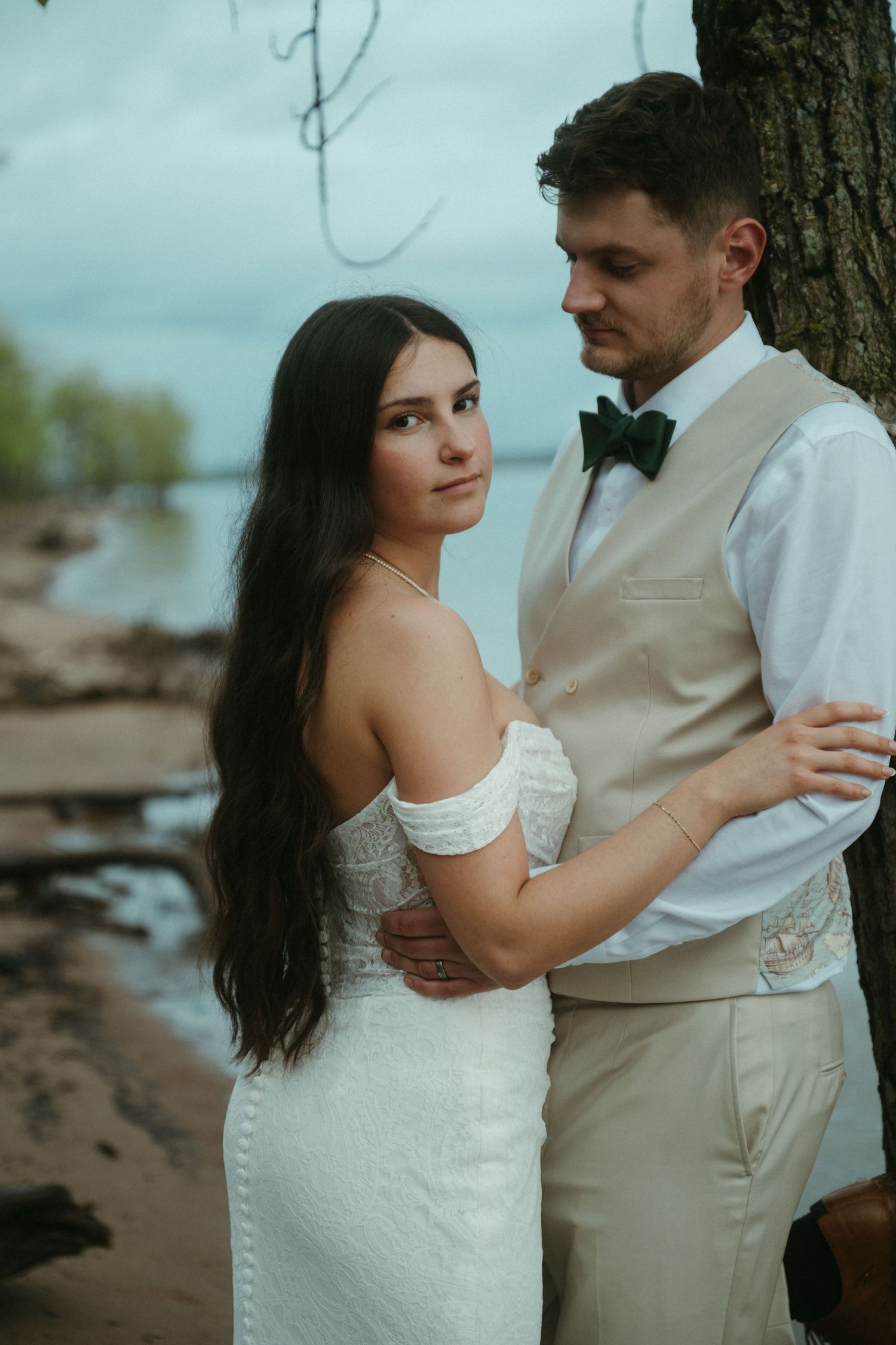 Bride in off-shoulder white gown embracing groom in beige vest and green bow tie by a lakeside tree.