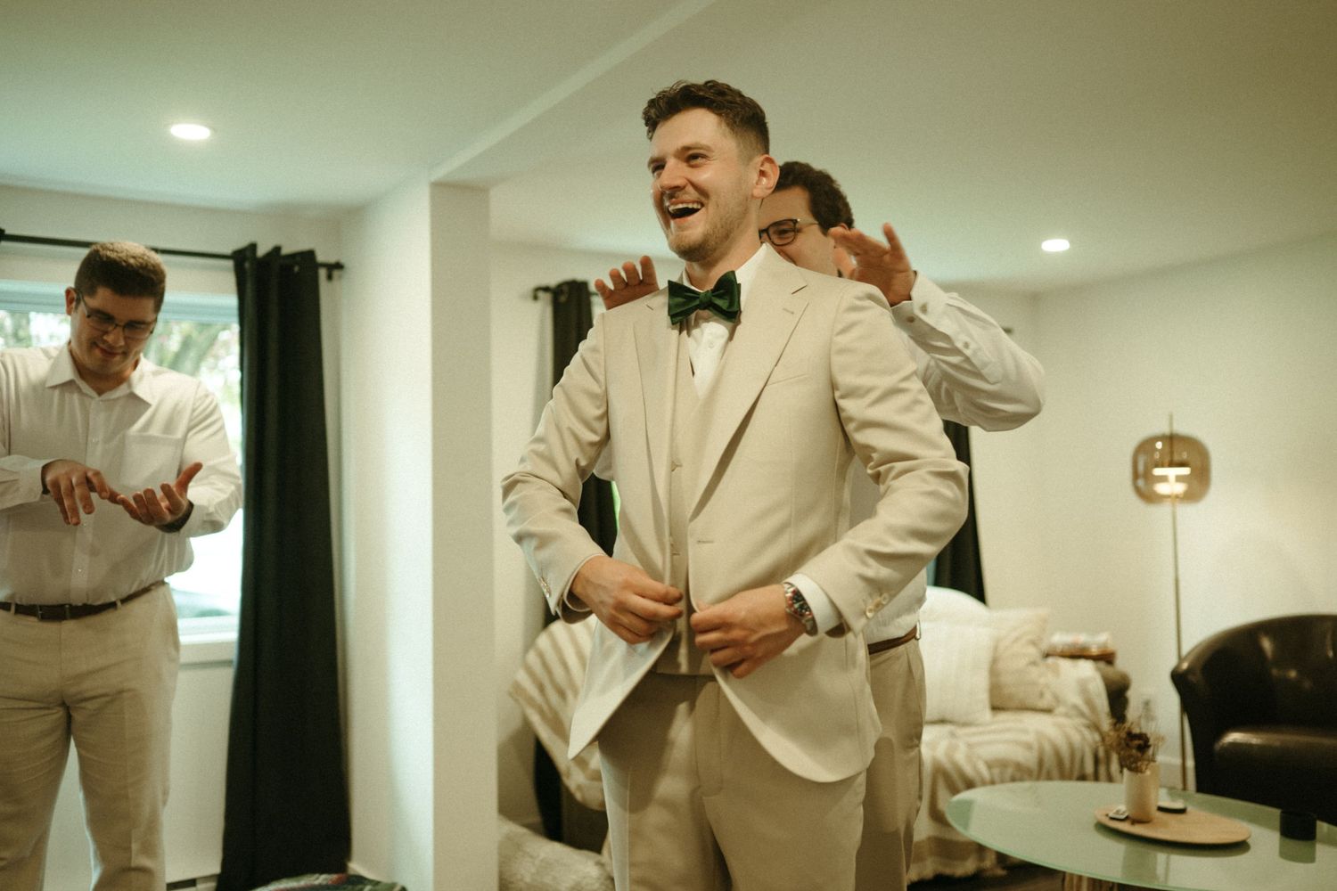 Groom laughing in beige suit with bow tie while groomsmen help him get ready.
