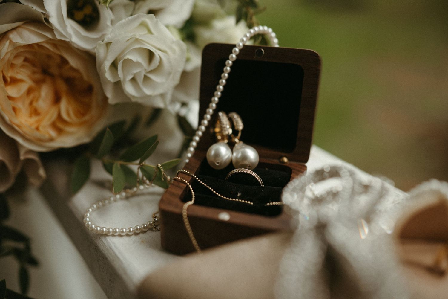 Elegant wedding jewelry flat lay featuring pearl earrings, a ring, and pearl necklace in a velvet box beside white roses.