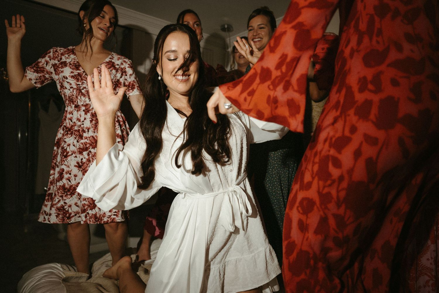 The image shows a group of women wearing colorful, floral-patterned dresses and posing together in a dimly lit setting.