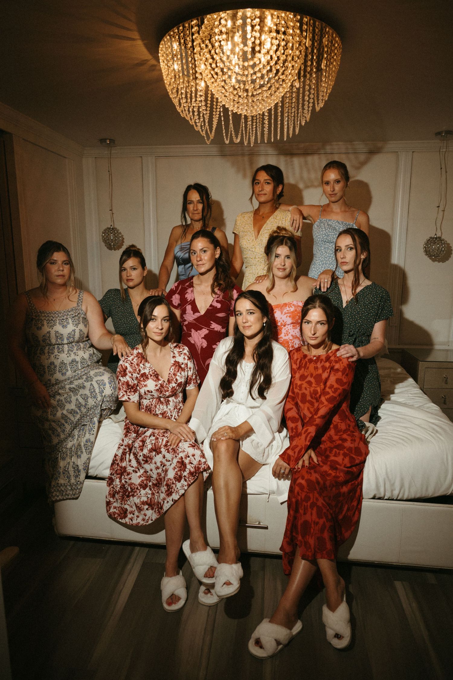 A group of women dressed in various colorful and patterned dresses and outfits, posing together in a room with a chandelier.
