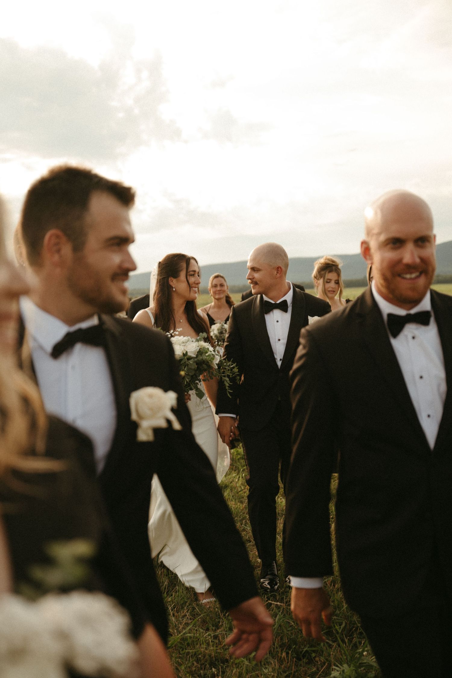 The image depicts a wedding party standing outdoors in a scenic, grassy field, with the couple in the center surrounded by their guests.