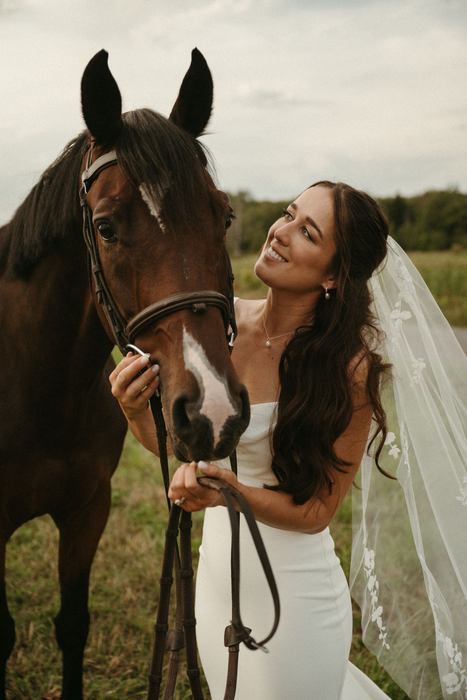 A woman in a white wedding dress stands next to a brown horse in a field.