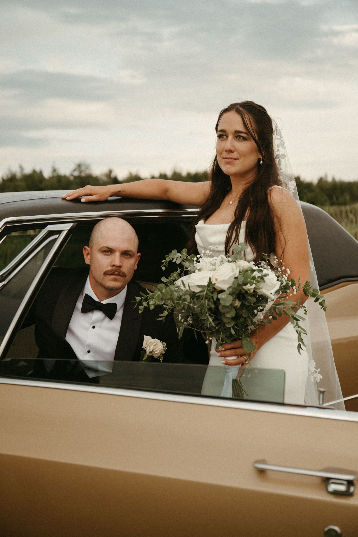A bride and groom sitting in a vintage car, the bride holding a bouquet of flowers.