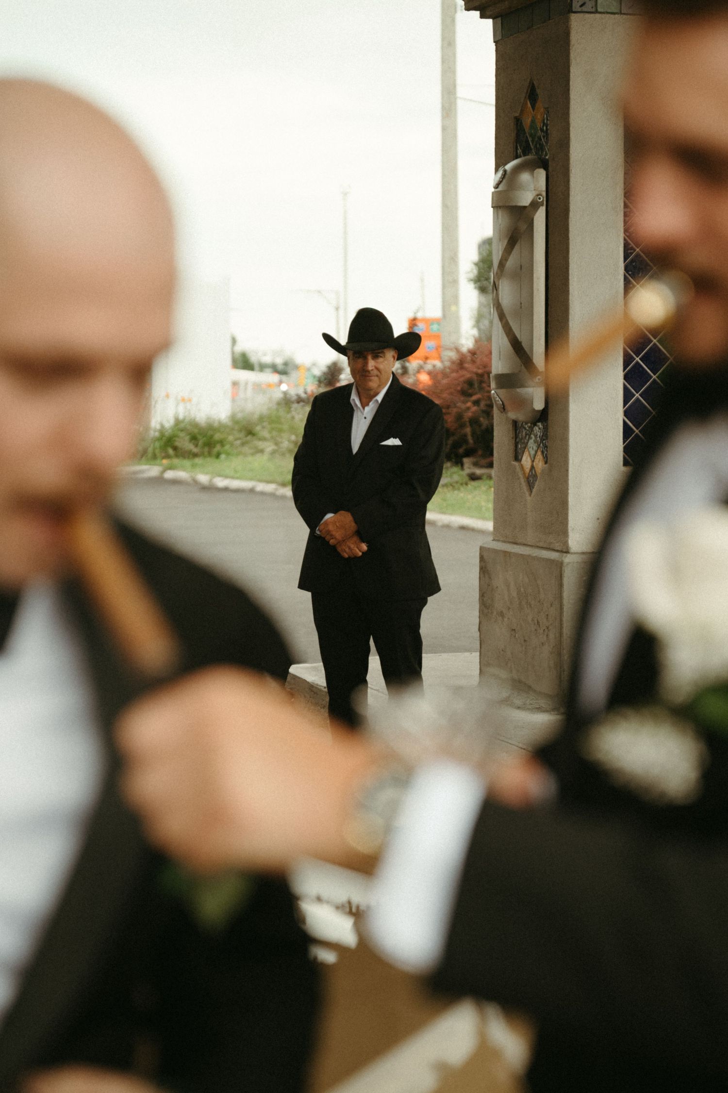 A man wearing a black suit and hat stands alone in an outdoor setting.