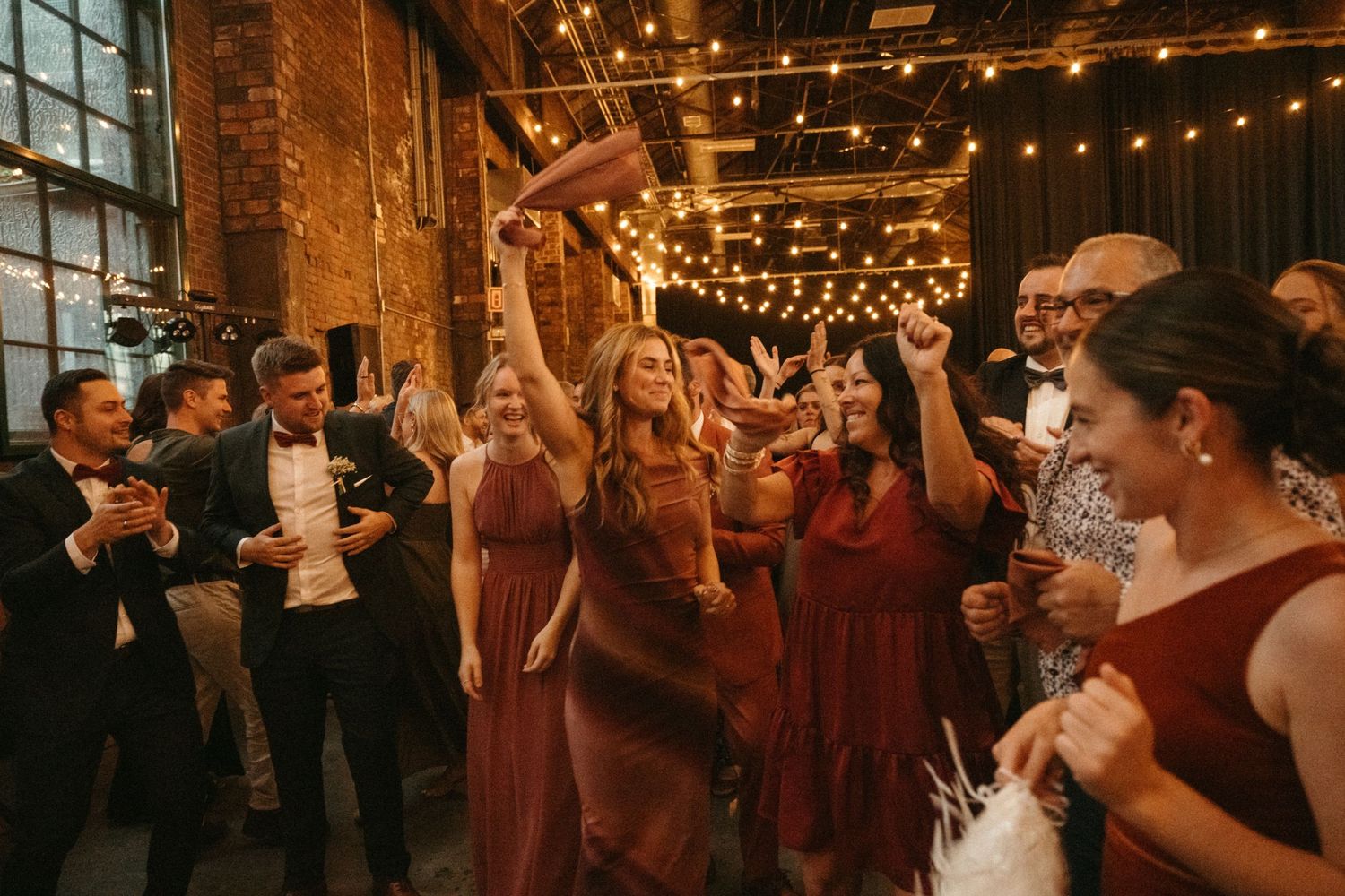 A group of people dancing and celebrating in a dimly lit, rustic indoor venue decorated with string lights.