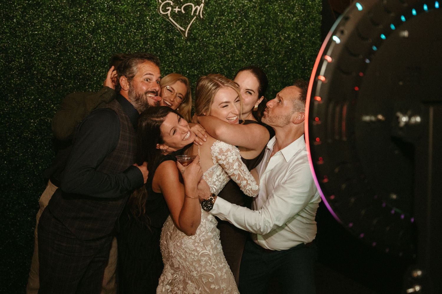 A group of people, including a bride in a white dress, are embracing and celebrating together at what appears to be a wedding reception.