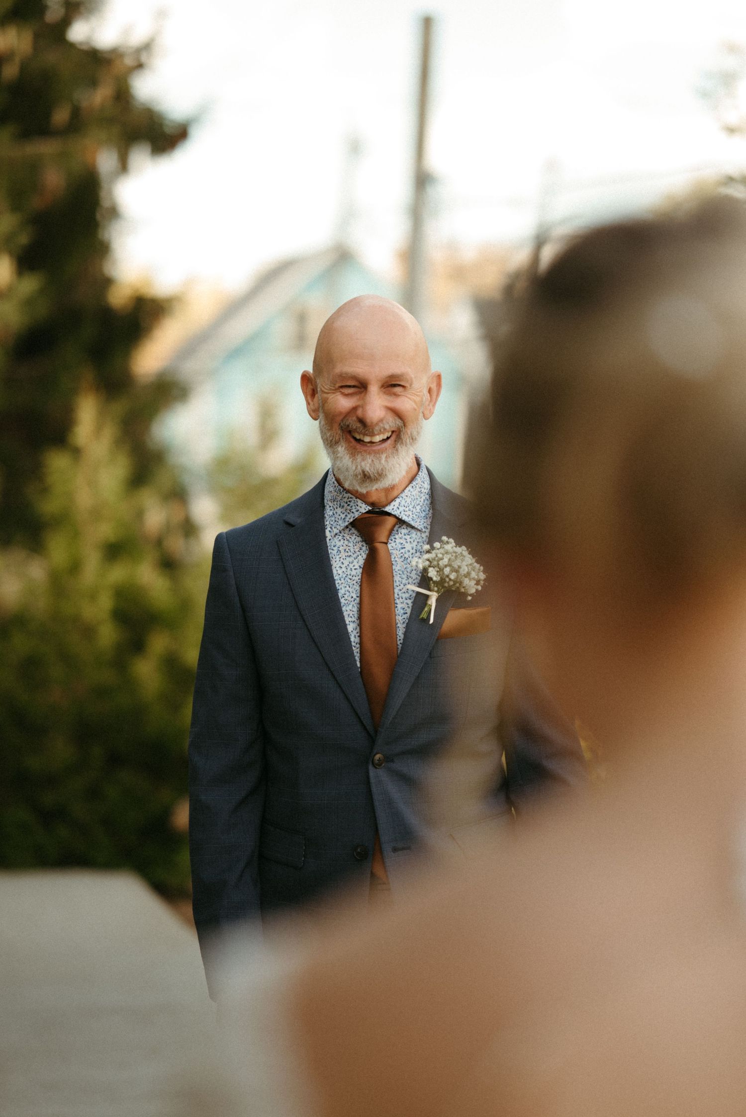 A smiling older man in a suit stands outdoors, surrounded by greenery.