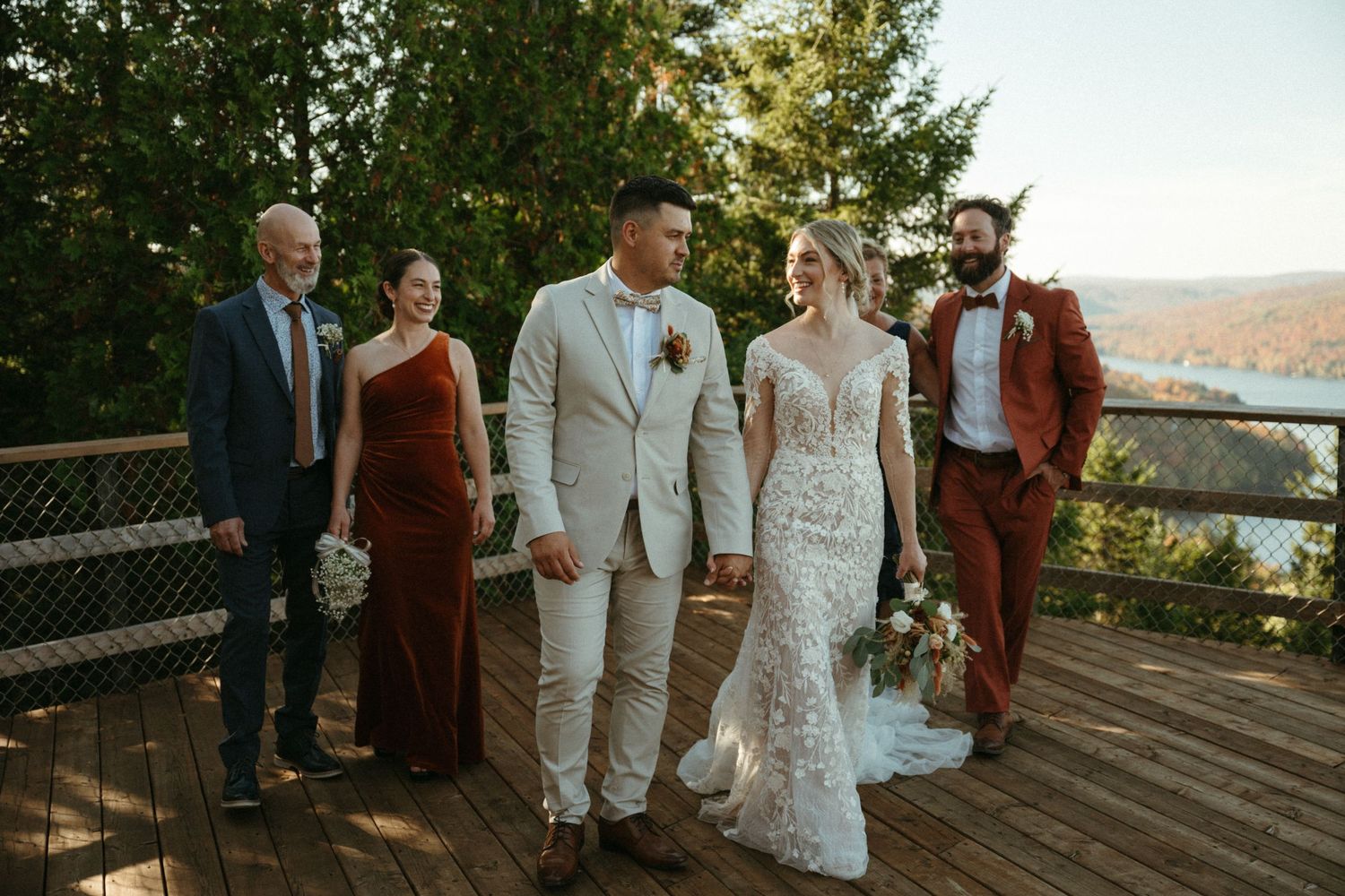 A group of people, including a bride and groom, stand on a wooden deck overlooking a scenic outdoor setting.