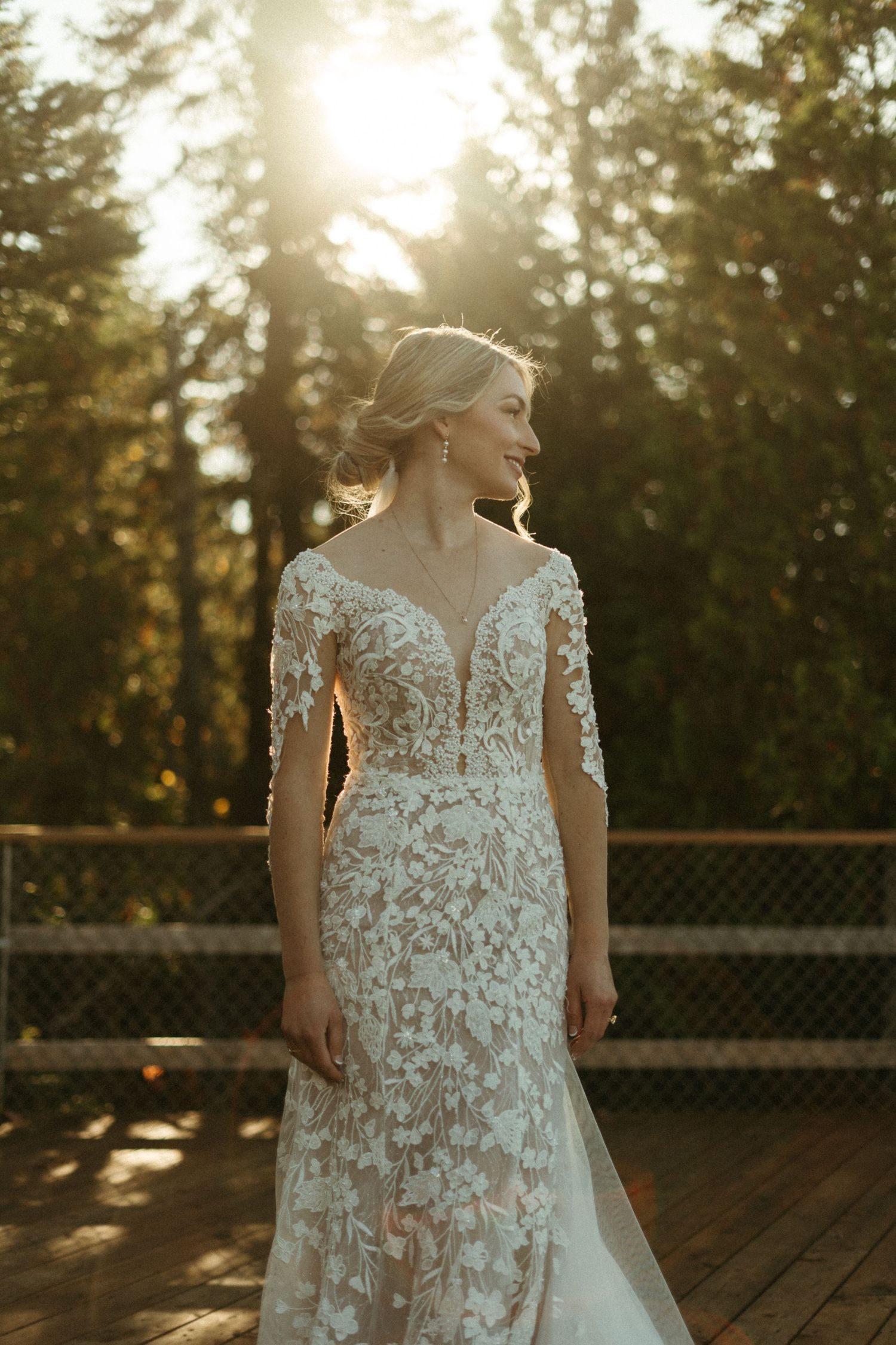 A woman in a lace wedding dress stands in a sunlit forest, her back to the camera.