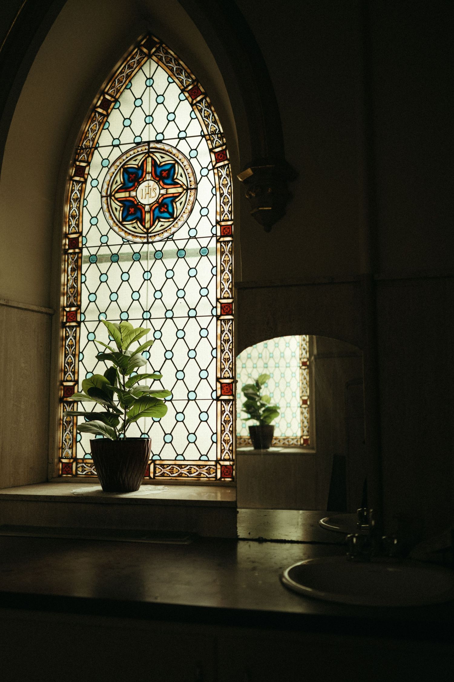 Stained glass Gothic church window with potted plants on stone sill beside a sink in dim light.