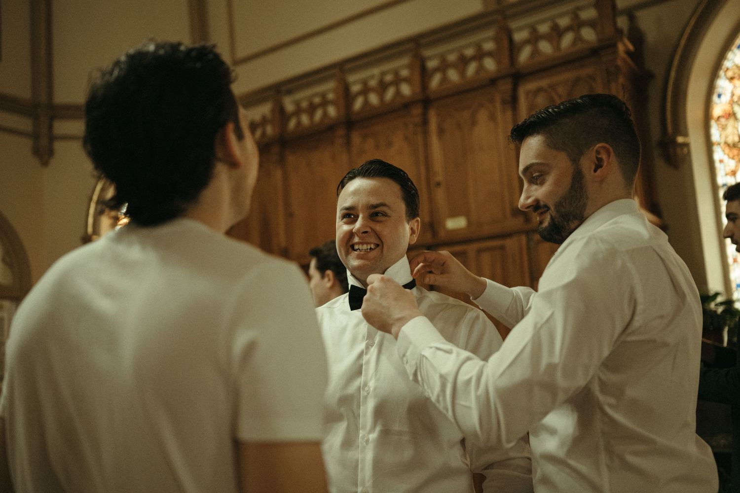 Groomsmen in white shirts helping groom adjust bow tie while laughing inside elegant church before wedding ceremony.