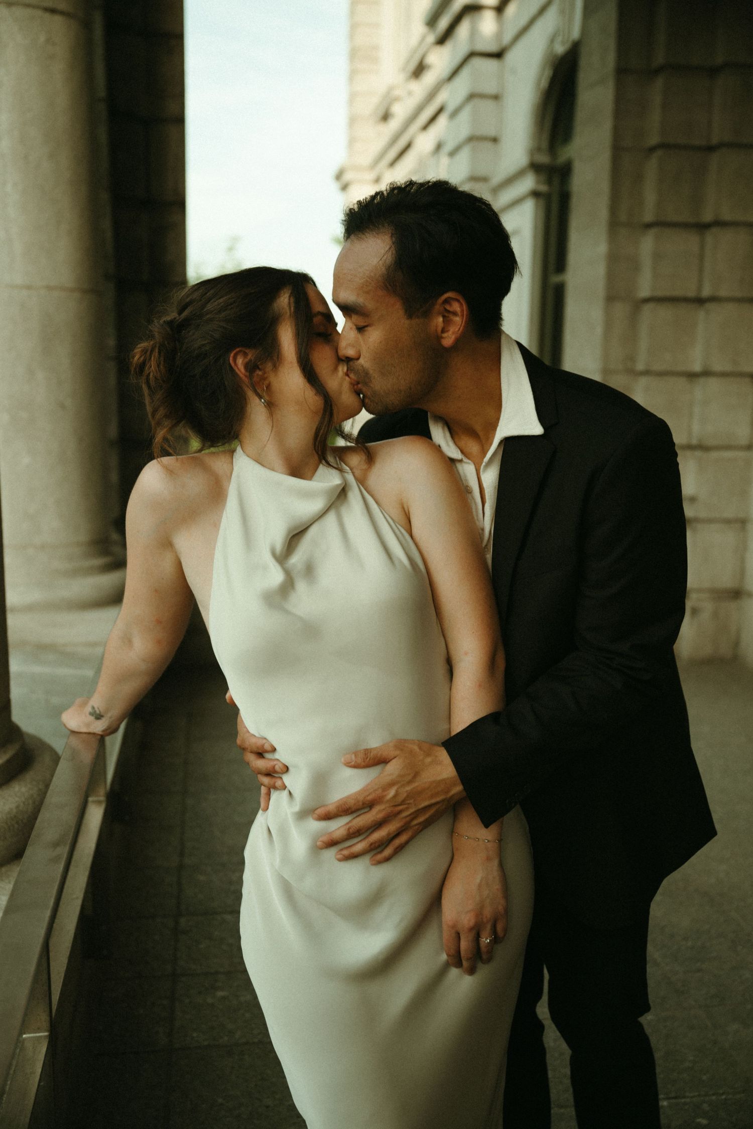 Elegant couple kissing in a neoclassical building corridor, bride in sleek white halter dress and groom in black suit.