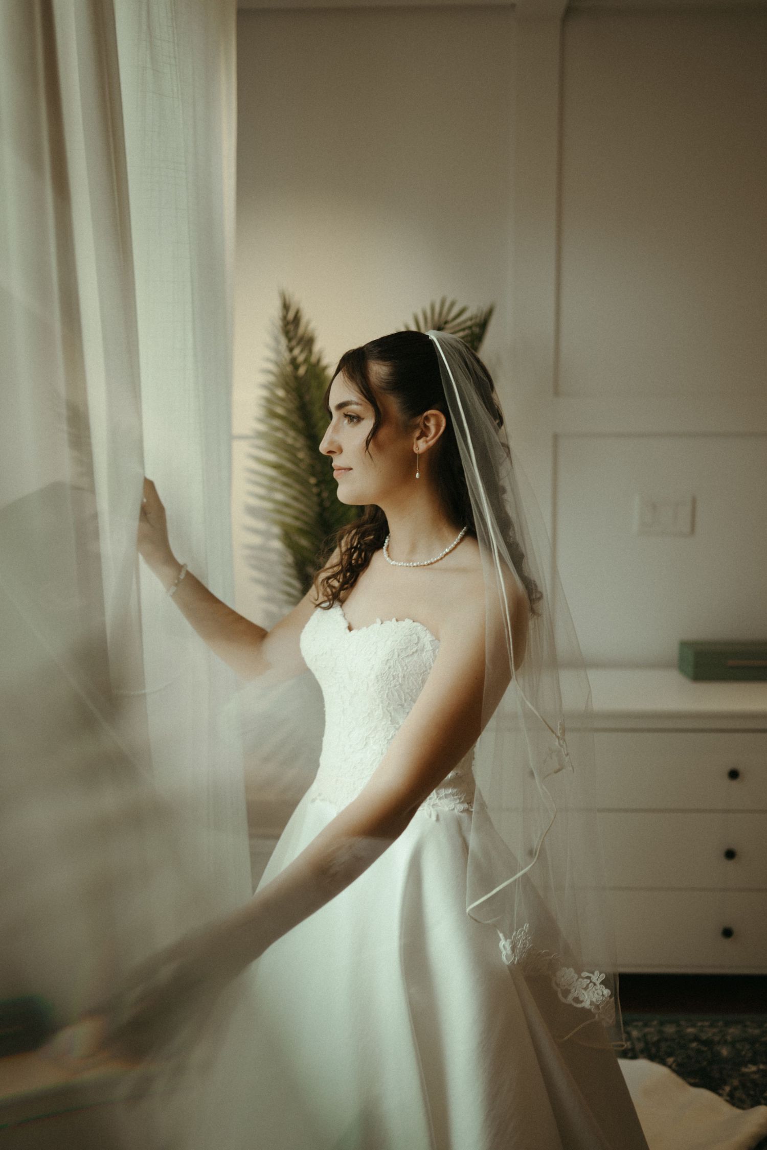 An elegant bride in a strapless white wedding gown and veil stands by a curtained window in soft light.