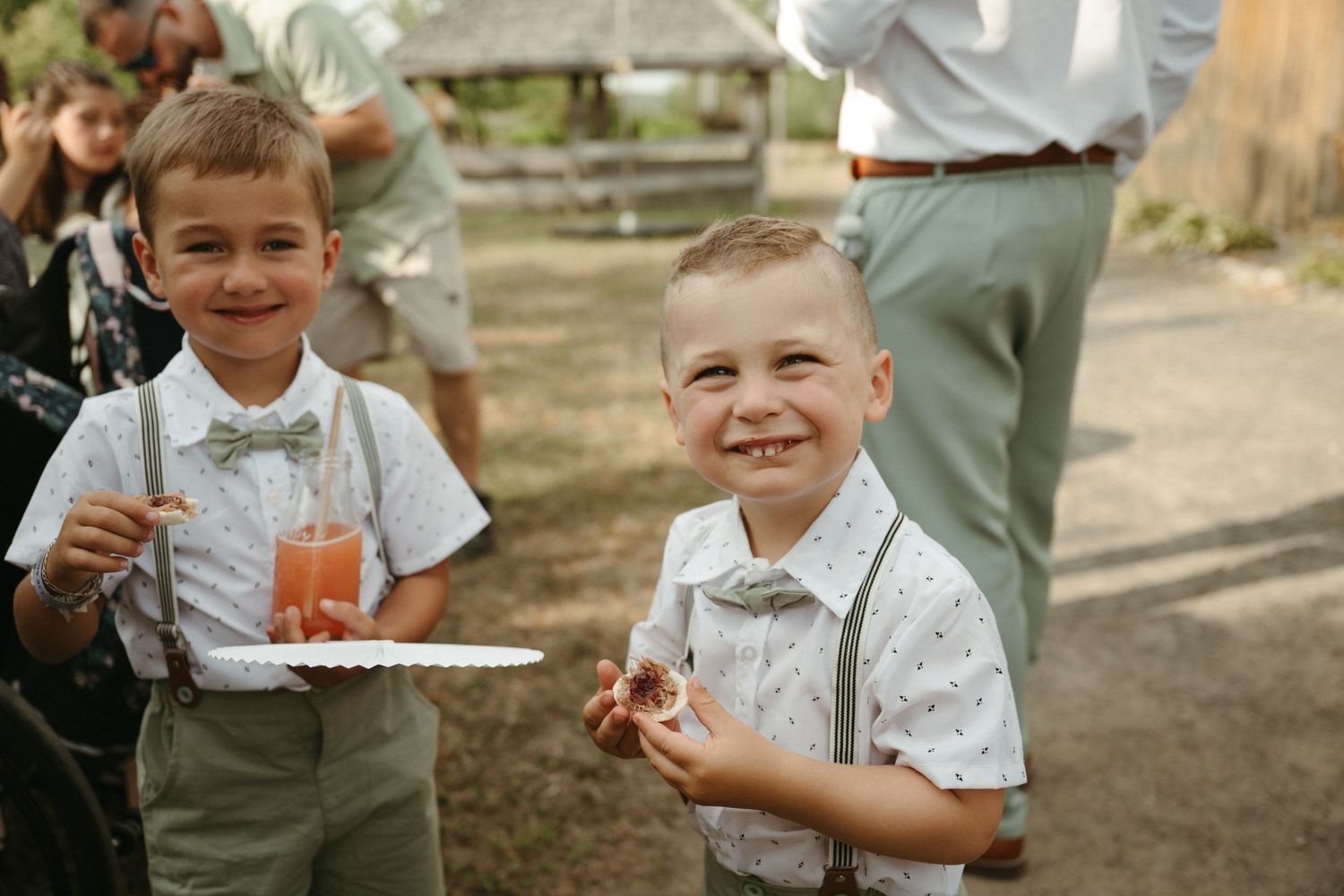 Two young ring bearers in suspenders and bow ties smile while enjoying food at an outdoor wedding.