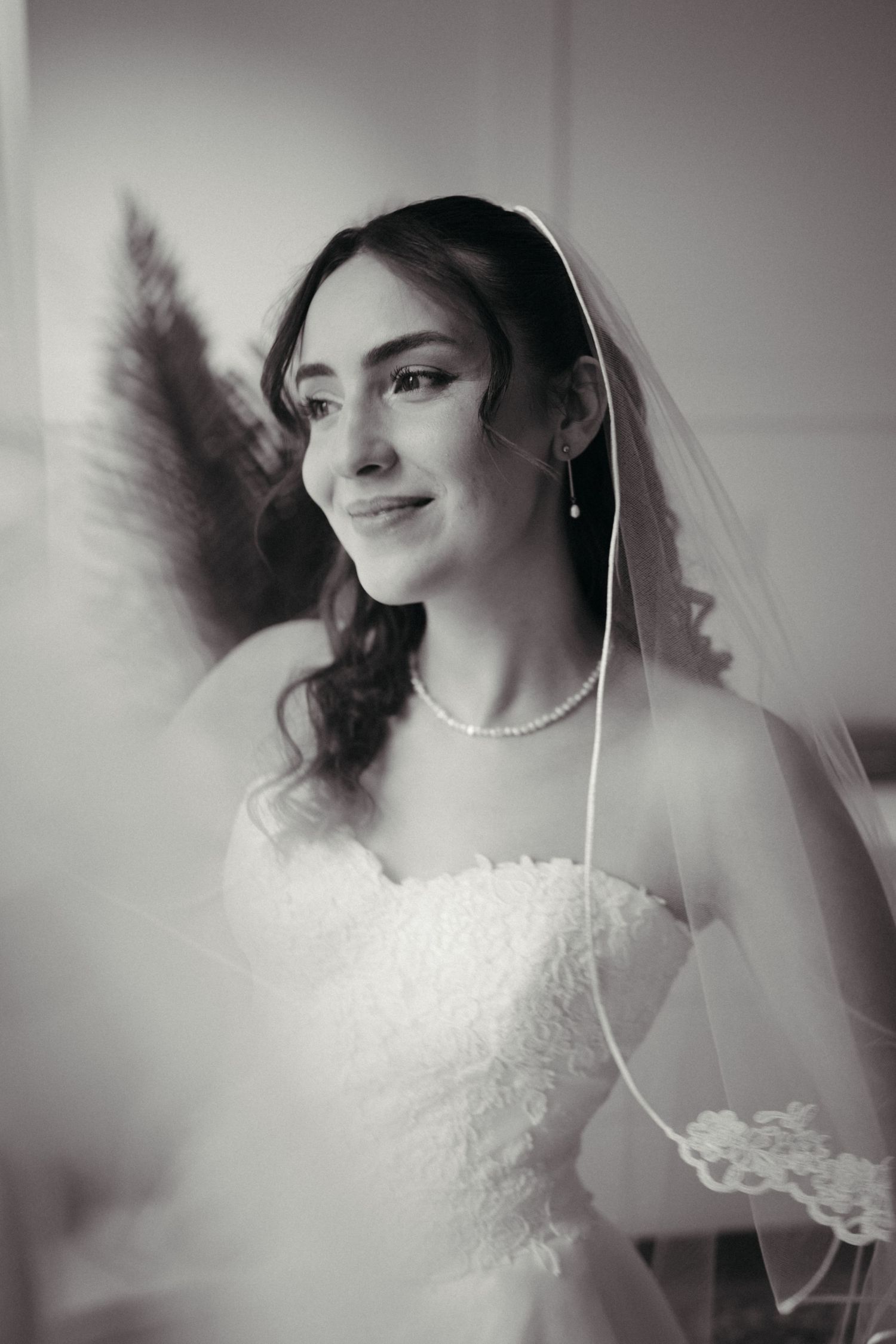 Black and white portrait of a bride in a lace dress, pearl necklace, and veil smiling softly near a window.