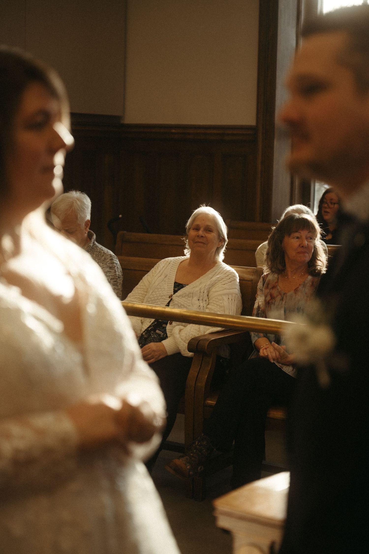 Wedding guests seated in wooden church pews watching a ceremony with bride and groom in foreground.