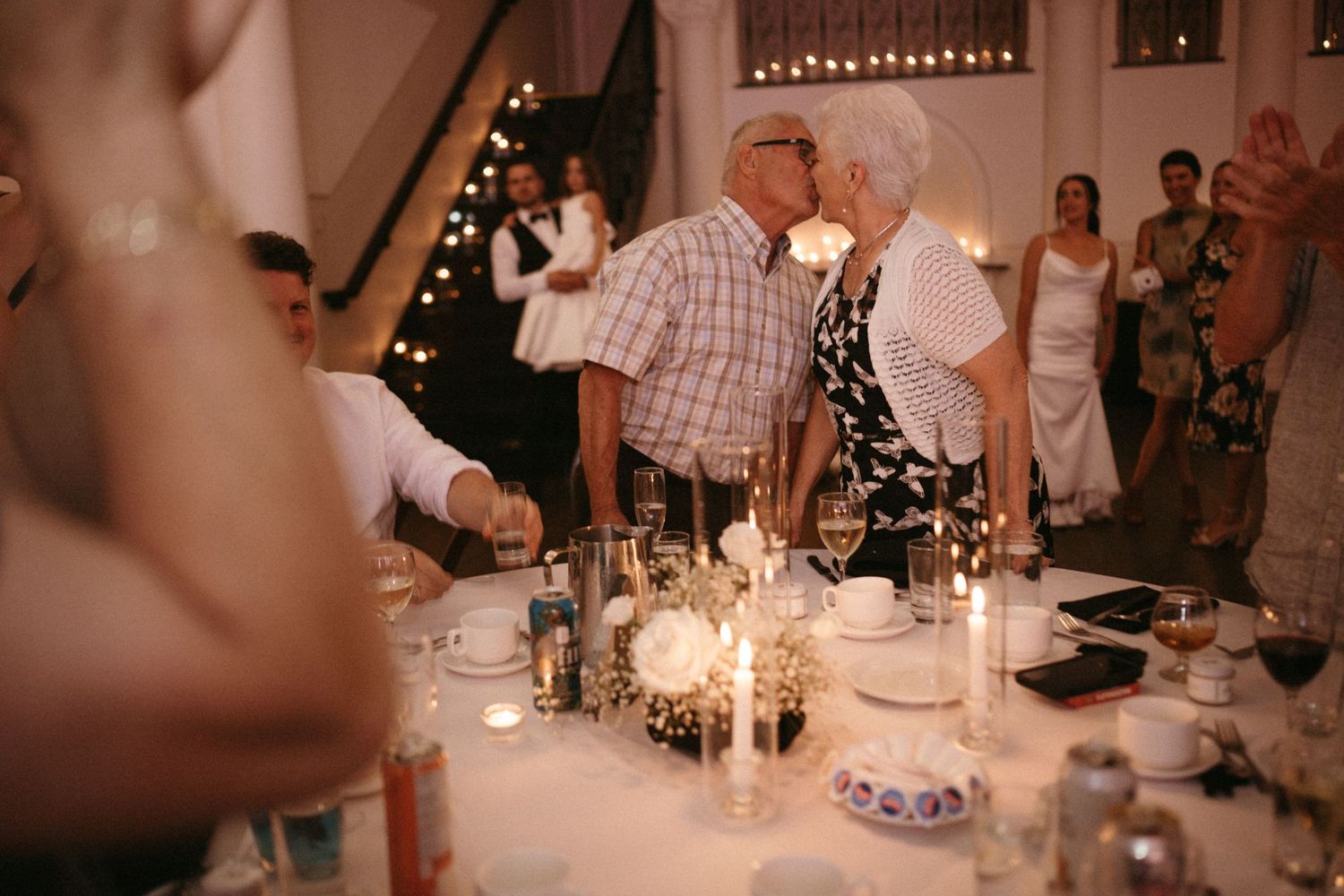 Elderly couple shares a sweet kiss at a candlelit wedding reception dinner table.