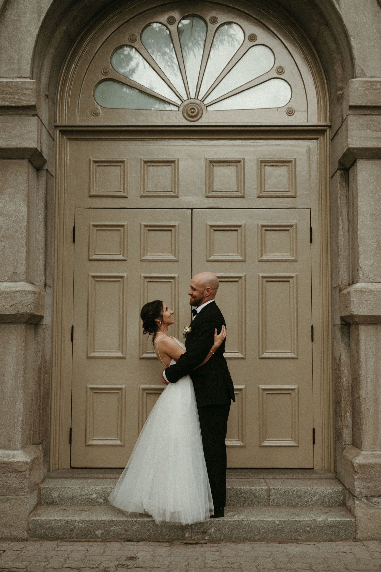 Bride and groom embracing romantically in front of elegant arched wooden doors at church entrance.