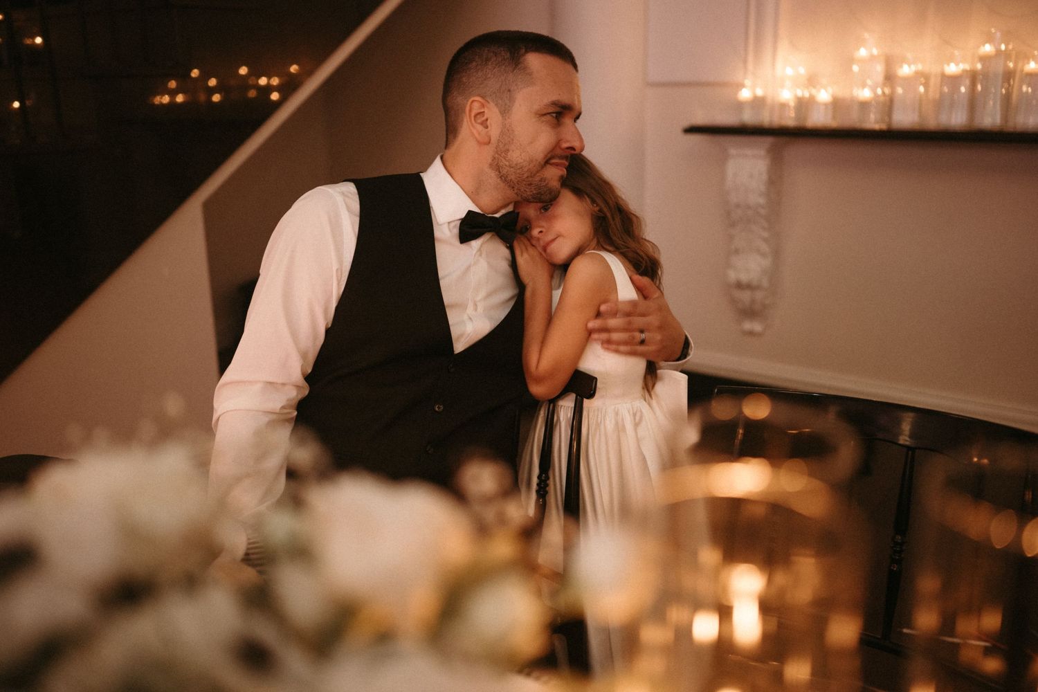 Groom in vest and bow tie tenderly embraces flower girl at candlelit wedding reception table.
