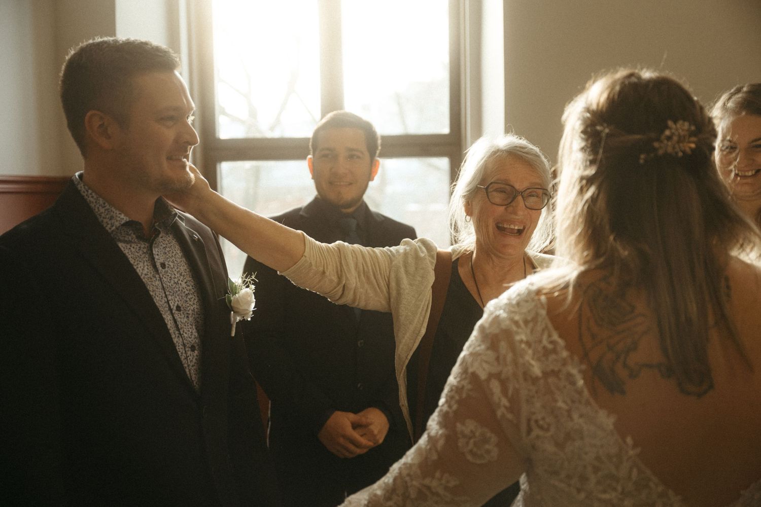 Joyful wedding moment with bride in lace gown, groom in suit, and guests celebrating indoors in warm light.