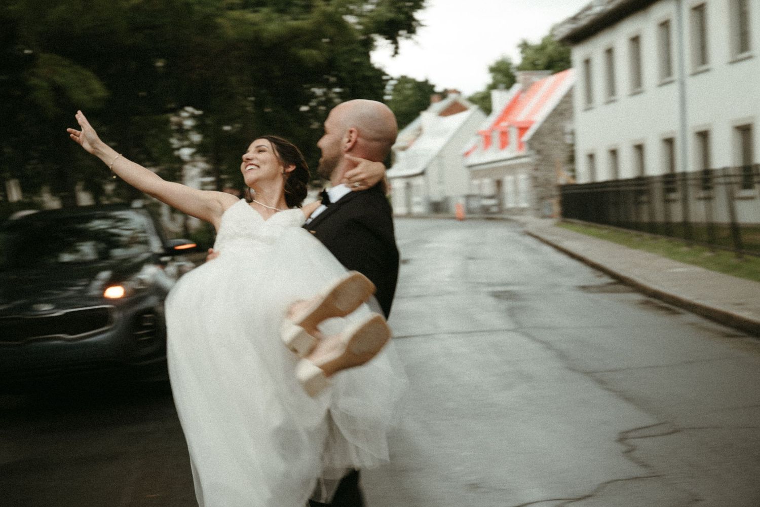 Groom carrying joyful bride in white ball gown through rainy street in romantic wedding photo.