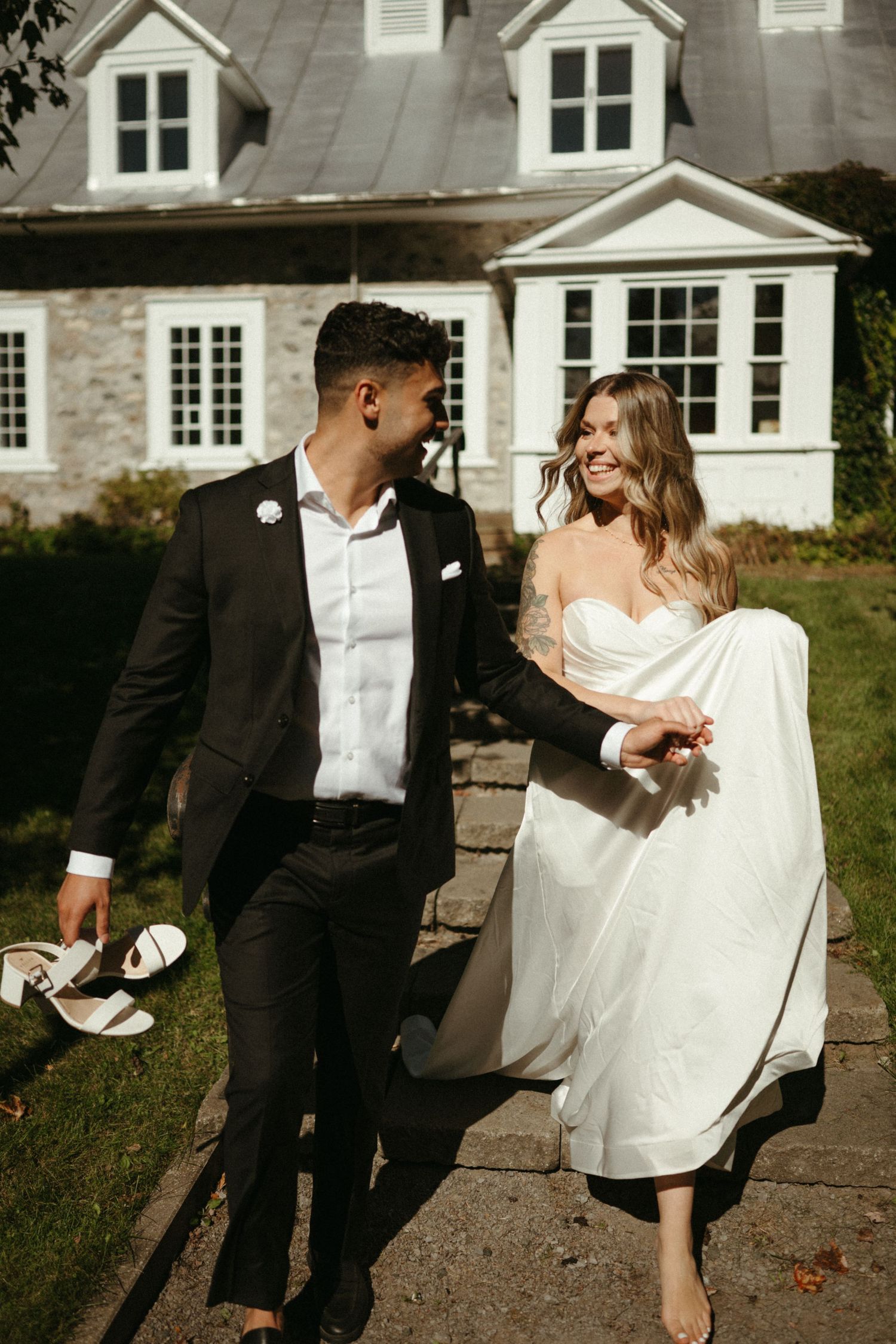 A well-dressed couple walks together on a path in front of a large, traditional-style house.