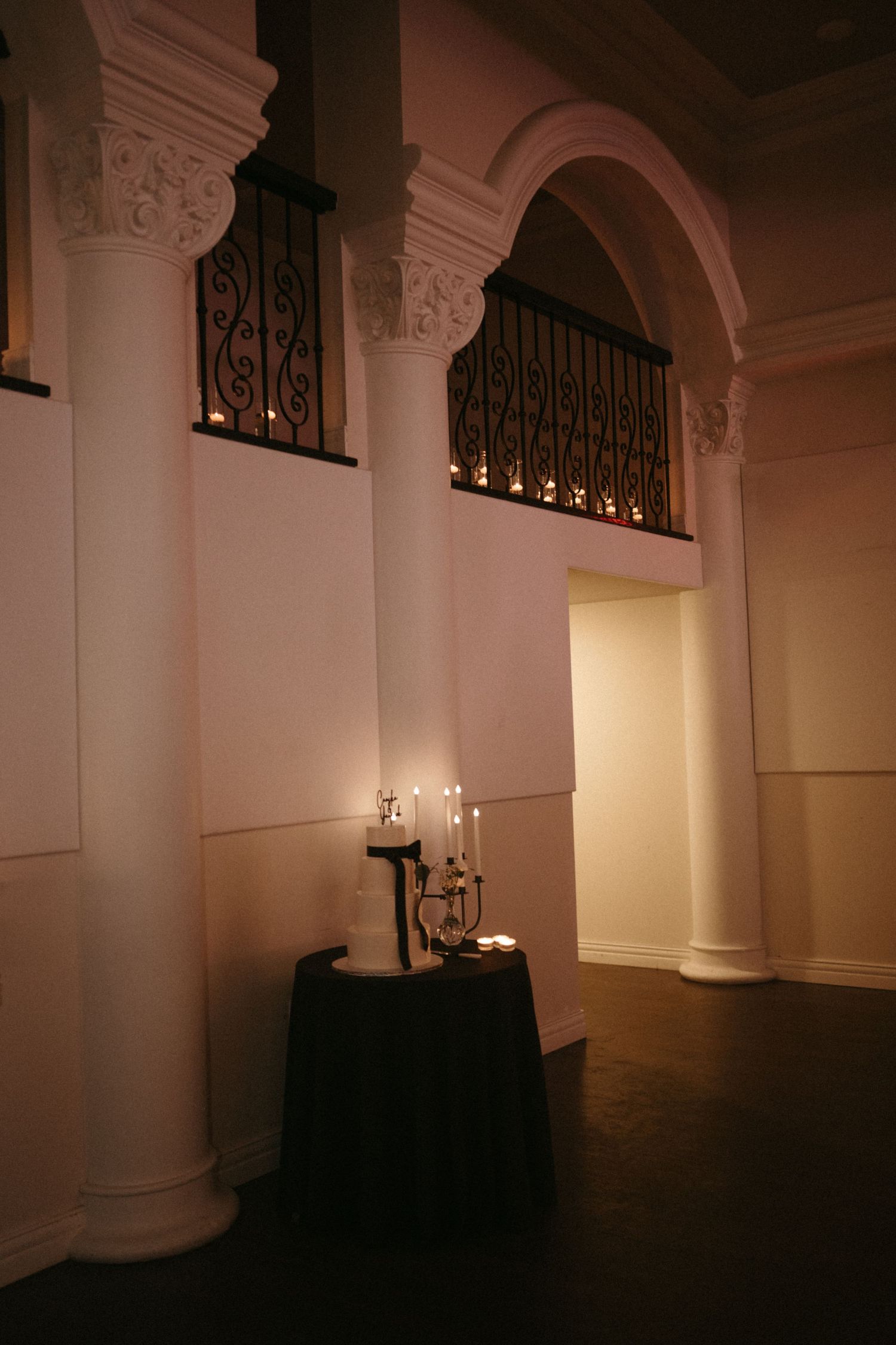Elegant wedding venue interior with white columns, wrought iron balcony, and candlelit decor in warm ambient lighting.
