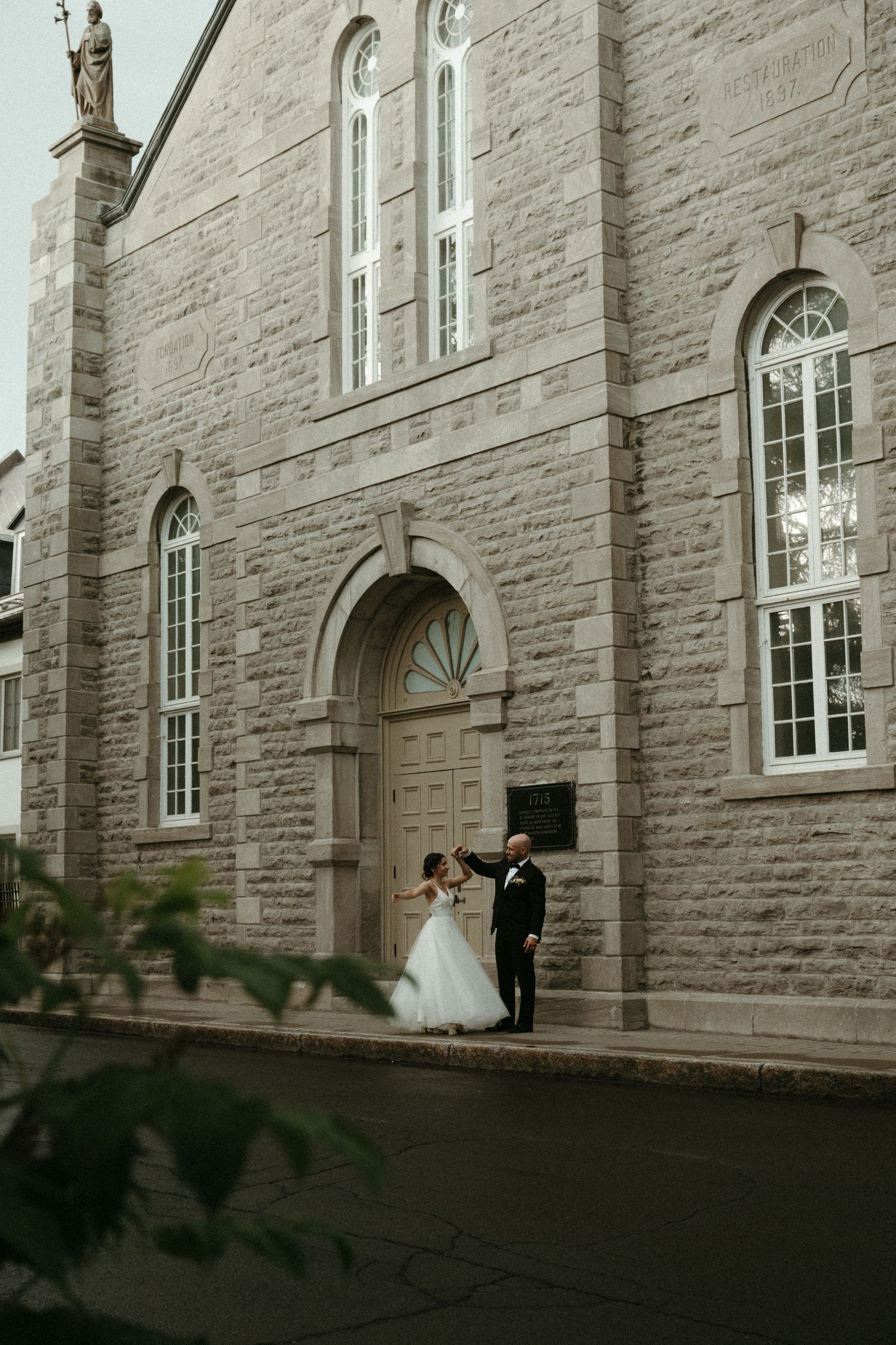 Bride and groom dancing in front of historic stone church with arched wooden doors.