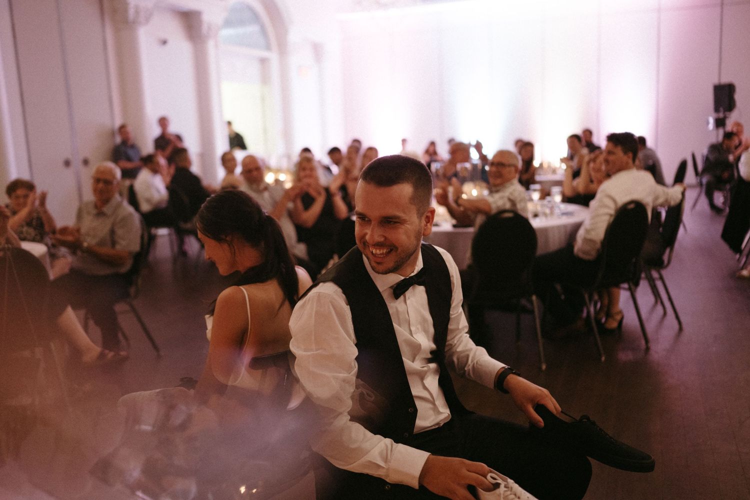 Laughing groom in black tuxedo vest enjoys wedding reception surrounded by seated guests.