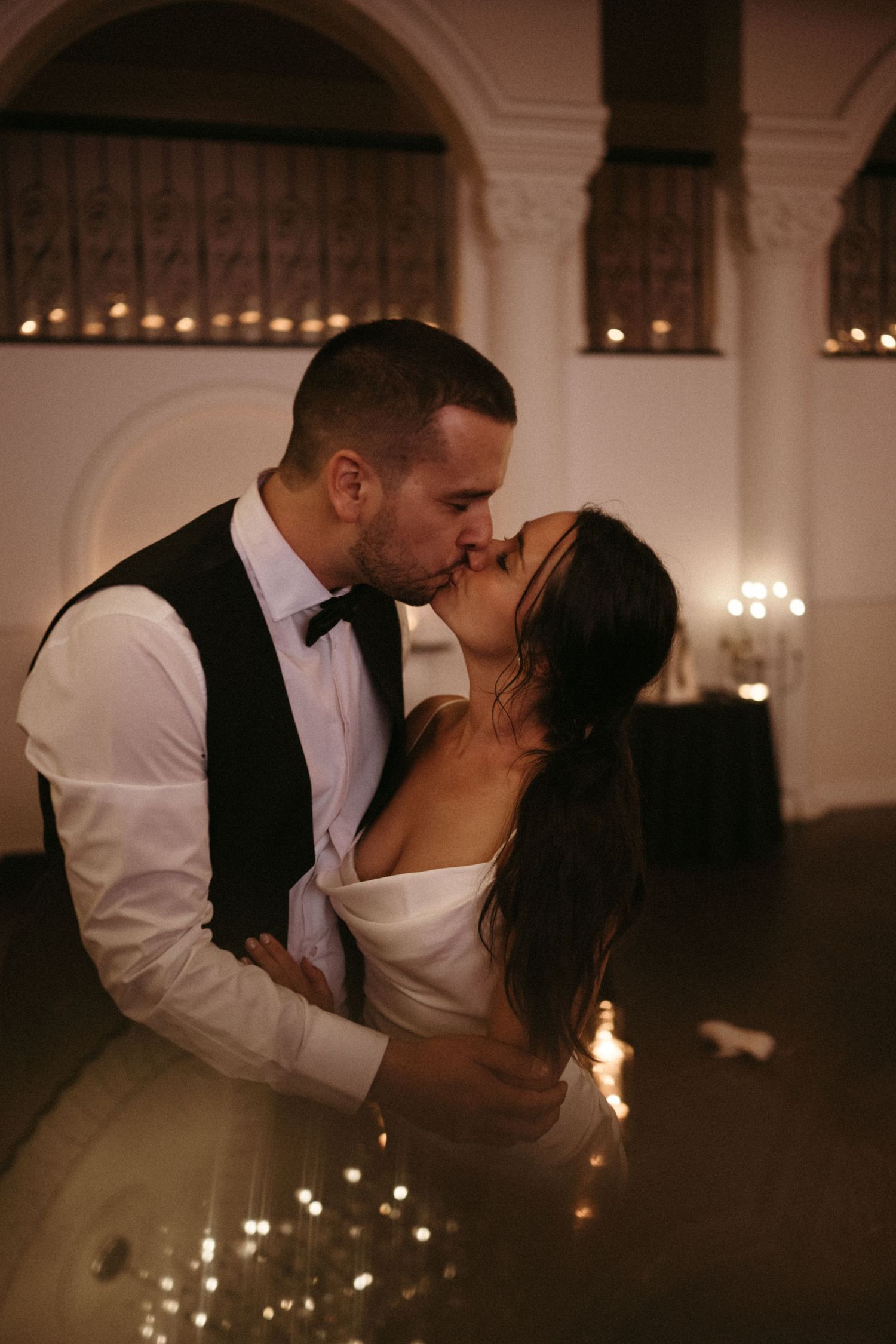 Newlyweds sharing passionate kiss on dance floor with sparkling candle lights in elegant wedding reception hall.