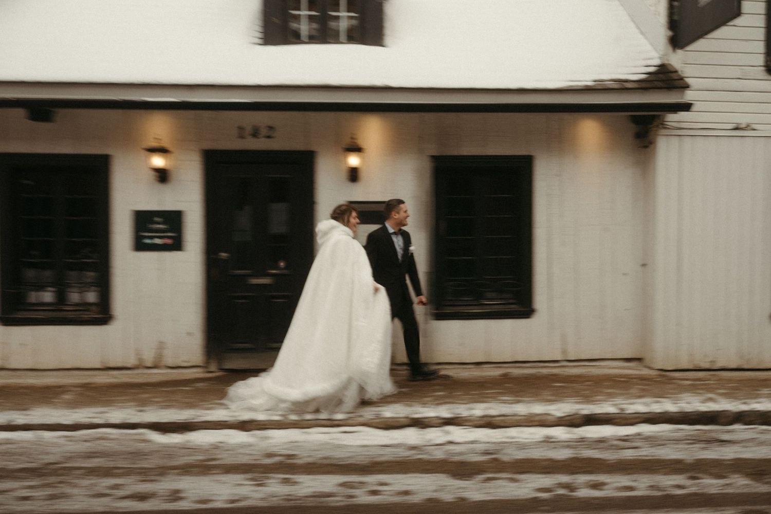 Bride in white gown and cape walks with groom in black tux outside snowy building 142.