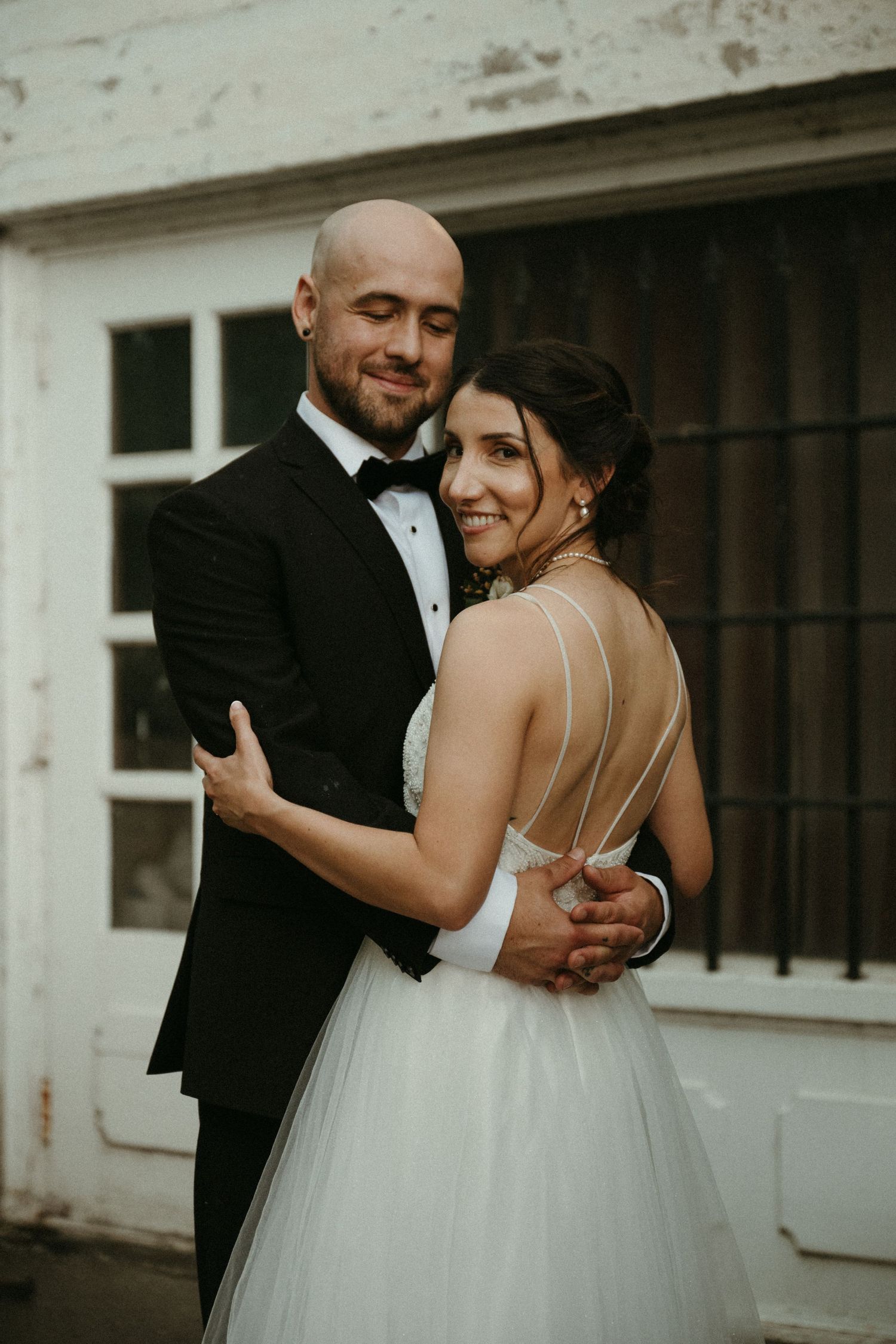 A bride in a backless white gown embraces her tuxedo-clad groom outdoors in a romantic wedding portrait.