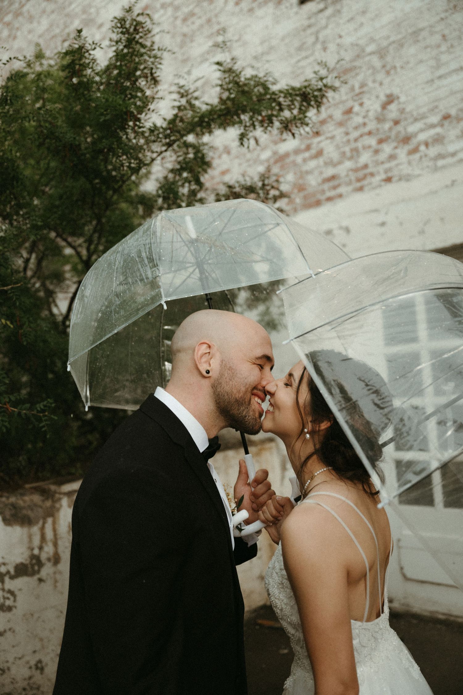 Bride and groom sharing romantic kiss under clear bubble umbrella against rustic brick wall backdrop.