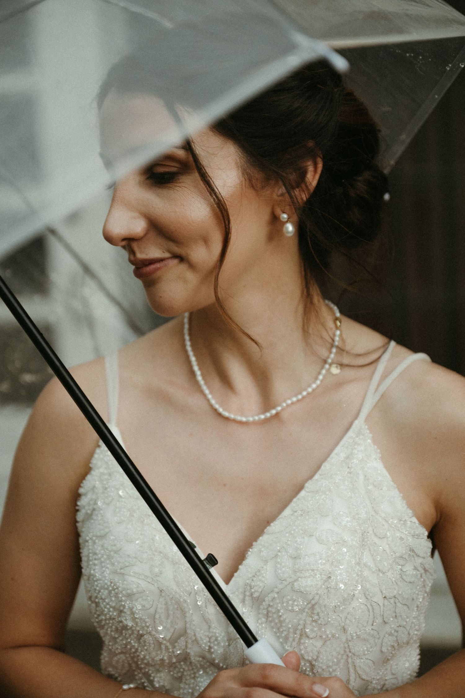 Elegant bride holding clear umbrella wearing pearl necklace and lace wedding gown.