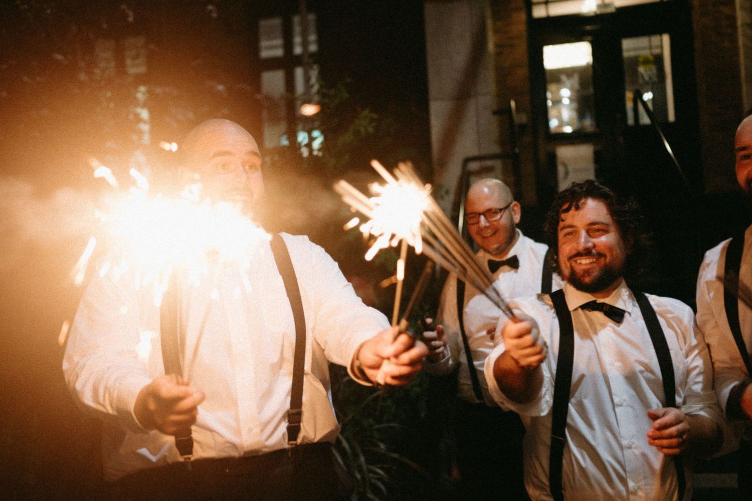 Men in suspenders and bow ties holding sparklers at a wedding reception celebration at night.