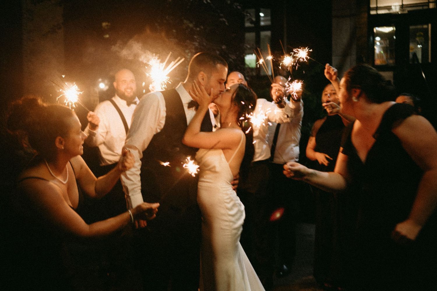 Bride and groom share a romantic kiss surrounded by wedding guests holding sparklers at a nighttime celebration.